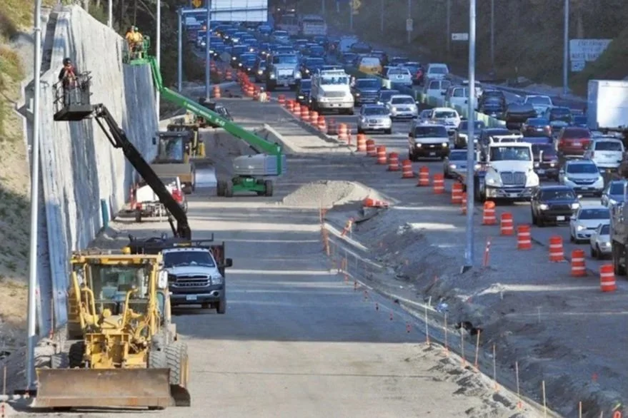 Road construction scene with construction vehicles and equipment on a highway, with traffic on the opposite side separated by orange traffic cones.