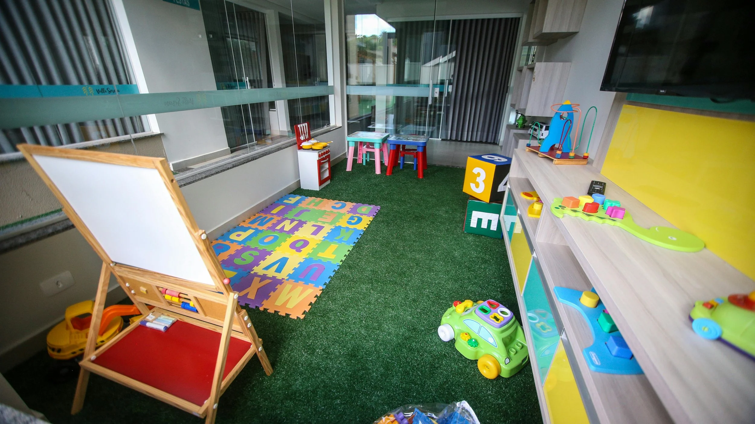 Indoor children's playroom with colorful toys, a foam alphabet puzzle mat, small tables and chairs, a whiteboard, and various educational toys and blocks on shelves.