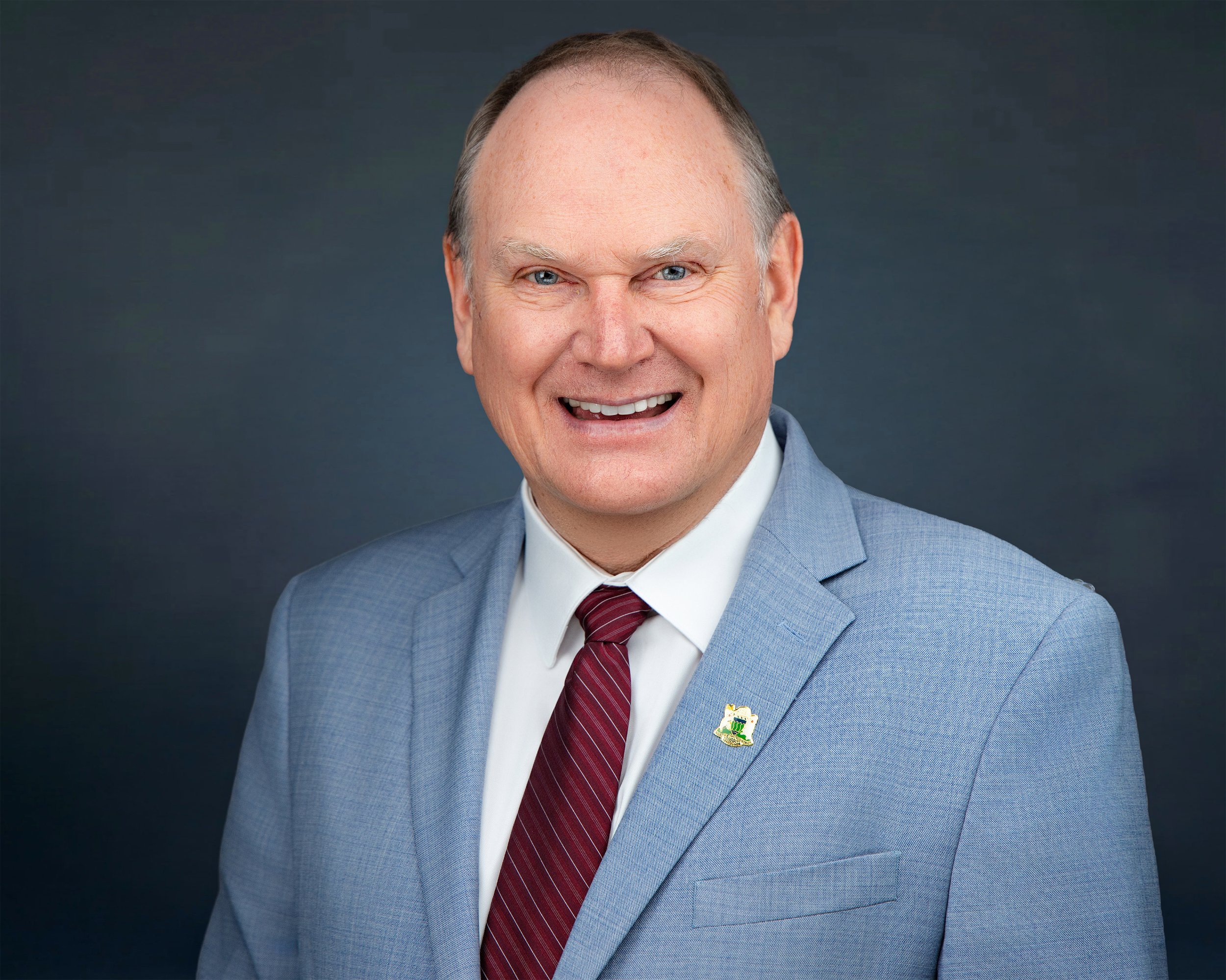 A smiling middle-aged man wearing a light blue blazer, white shirt, and a maroon tie, with a pin on his blazer's lapel.