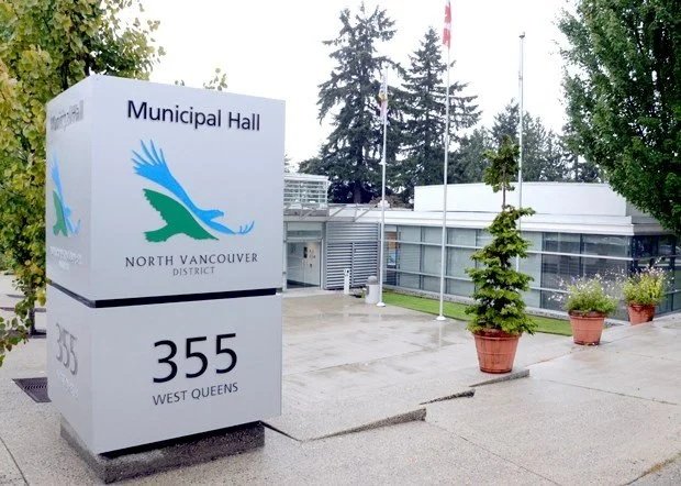Sign indicating the location of the North Vancouver District Municipal Hall at 355 West Queens, with potted plants and trees nearby.