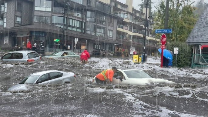 A street heavily flooded with water, with several cars partially submerged and people walking through the water. Buildings and street signs are visible in the background.