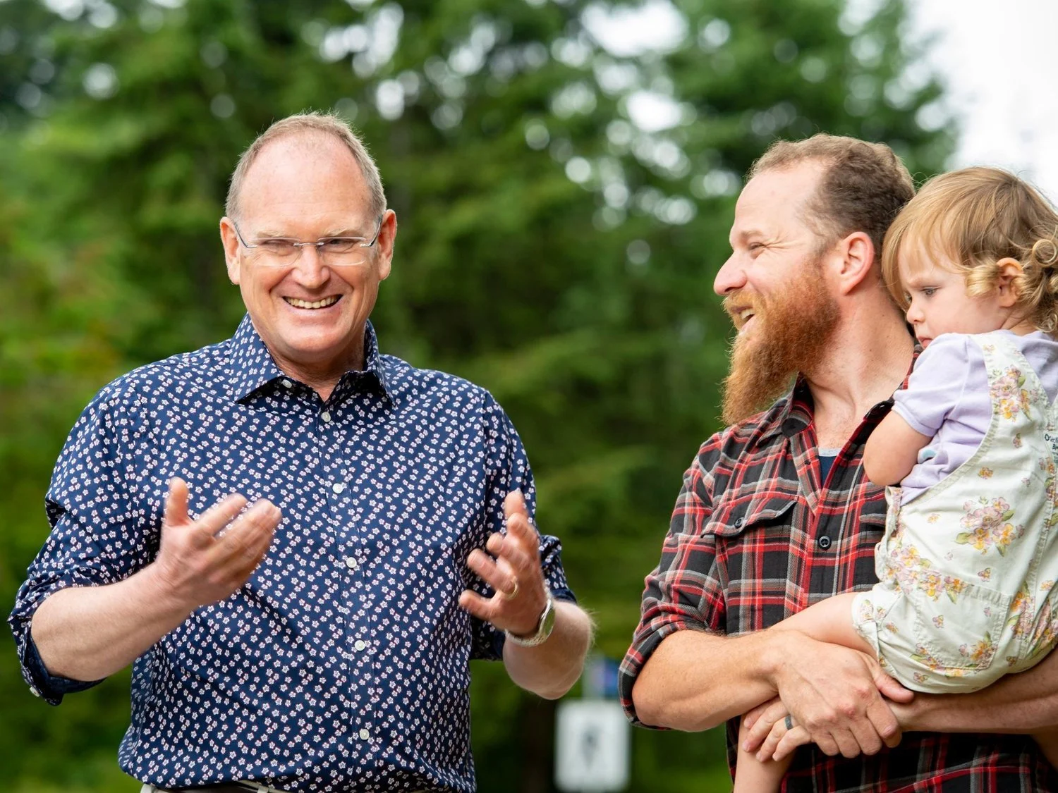 Two men standing outside, engaging in conversation; one is gesturing with his hands, smiling, and wearing glasses and a patterned shirt, while the other, with a beard and playful smile, is holding a young girl with curly hair in a light-colored dress, and they are surrounded by greenery.