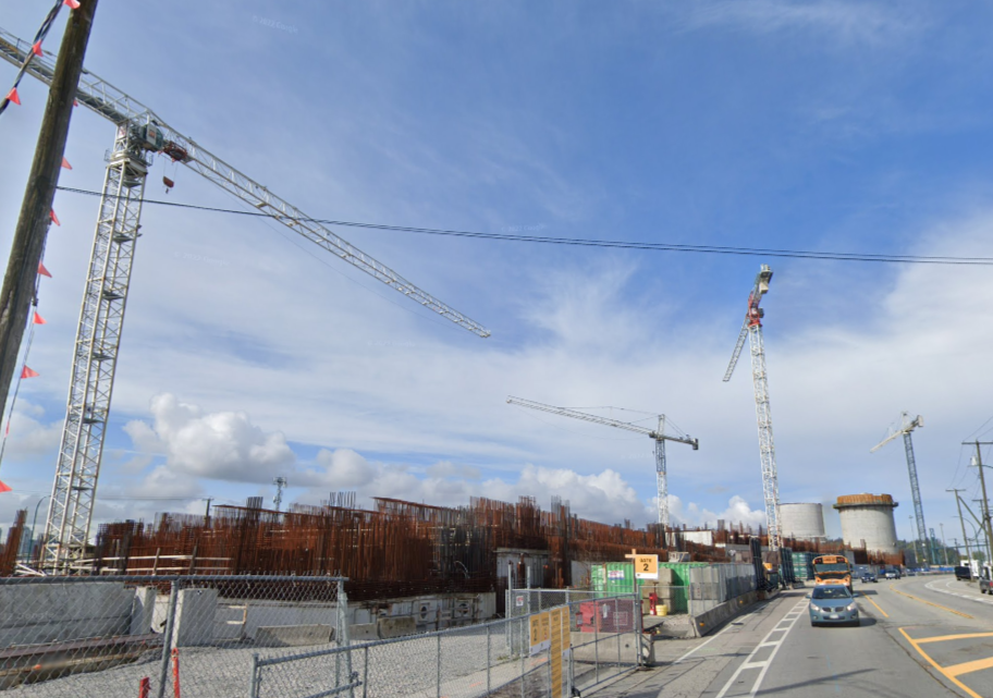 Construction site with multiple cranes, building structures, and construction equipment, alongside a road with cars under a partly cloudy sky.