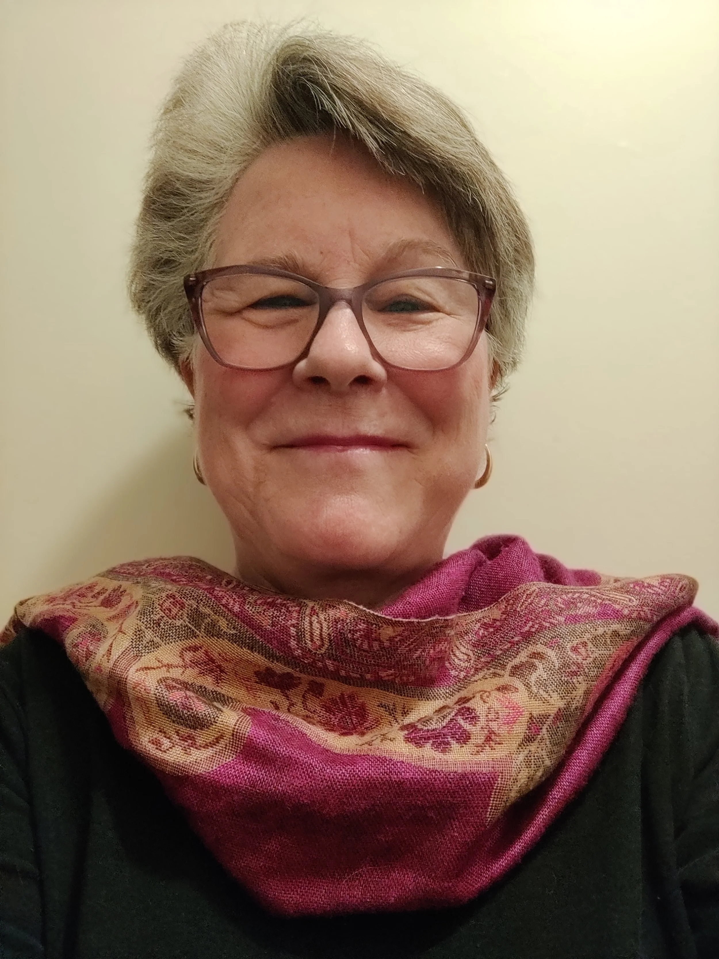 A smiling woman with short gray hair wearing glasses, a pink and beige patterned scarf, and a dark top, standing against a plain light-colored wall.