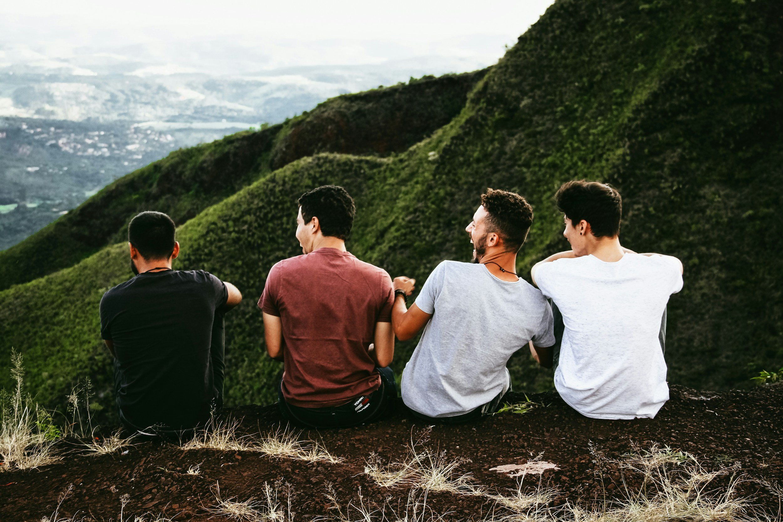 Four friends sitting on a hillside overlooking lush green mountains and a distant cityscape, engaged in conversation and enjoying the scenic view.