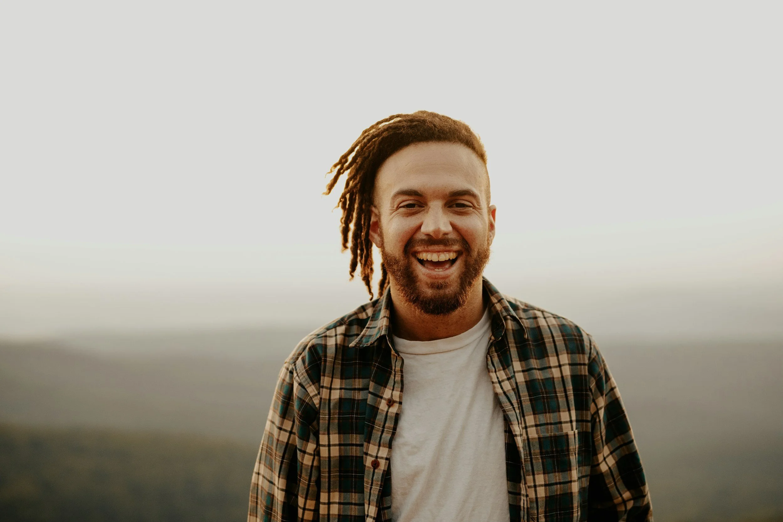A smiling man with dreadlocks and a beard standing outdoors during daytime, wearing a plaid shirt over a white t-shirt.