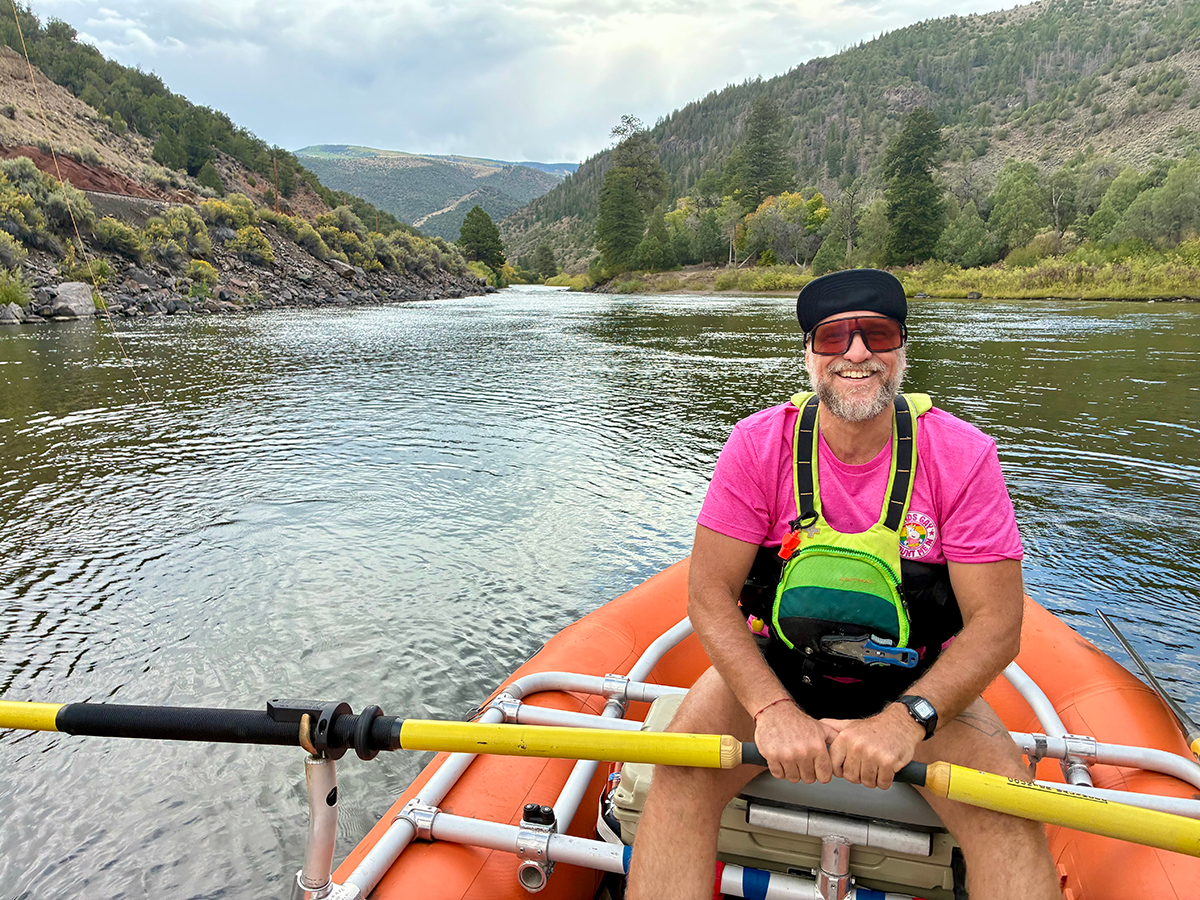 Smiling man in pink shirt and cap rowing on a river in inflatable boat with scenic mountains and trees in the background.