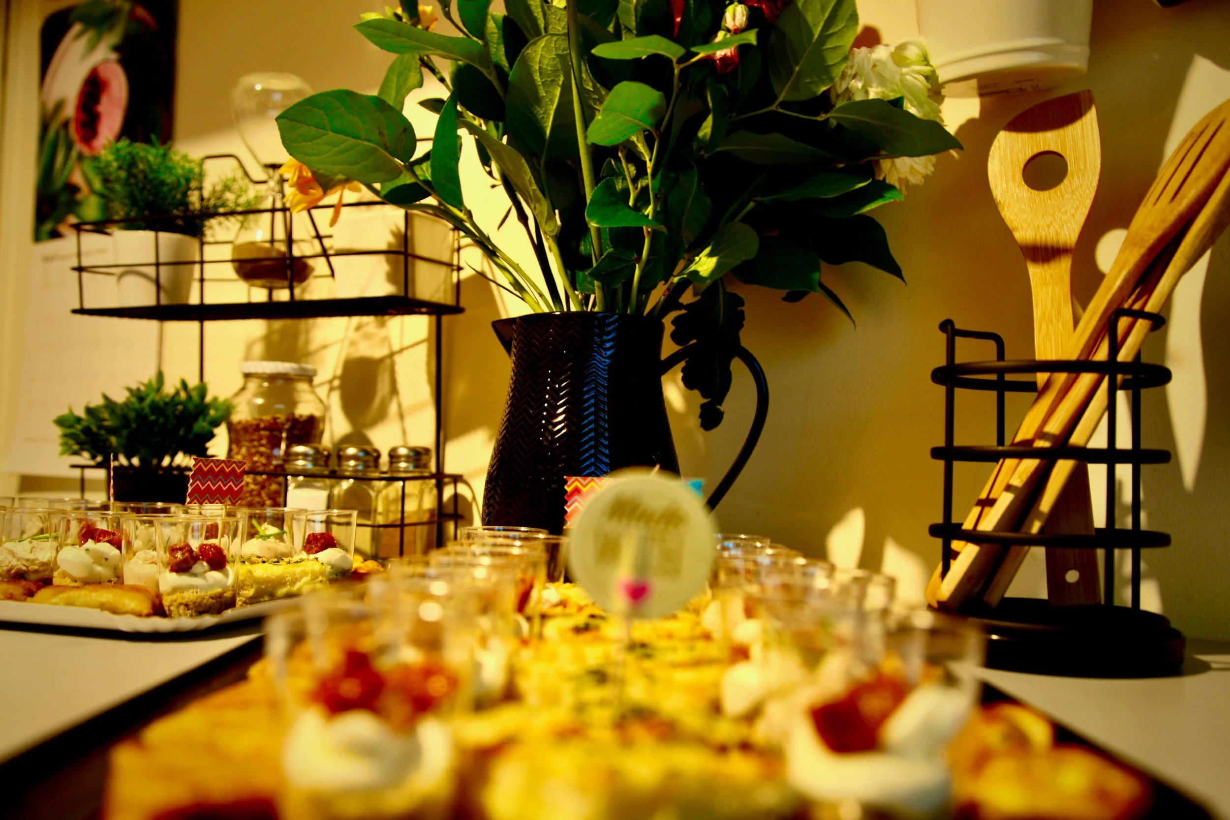 A table with an assortment of desserts, including small glasses with creamy treats topped with berries, arranged on a tray. Behind the desserts, there is a large black vase filled with green leafy plants. To the right, a wooden cutting board and tools are visible, and in the background, decorative items and potted plants are on a metallic rack.