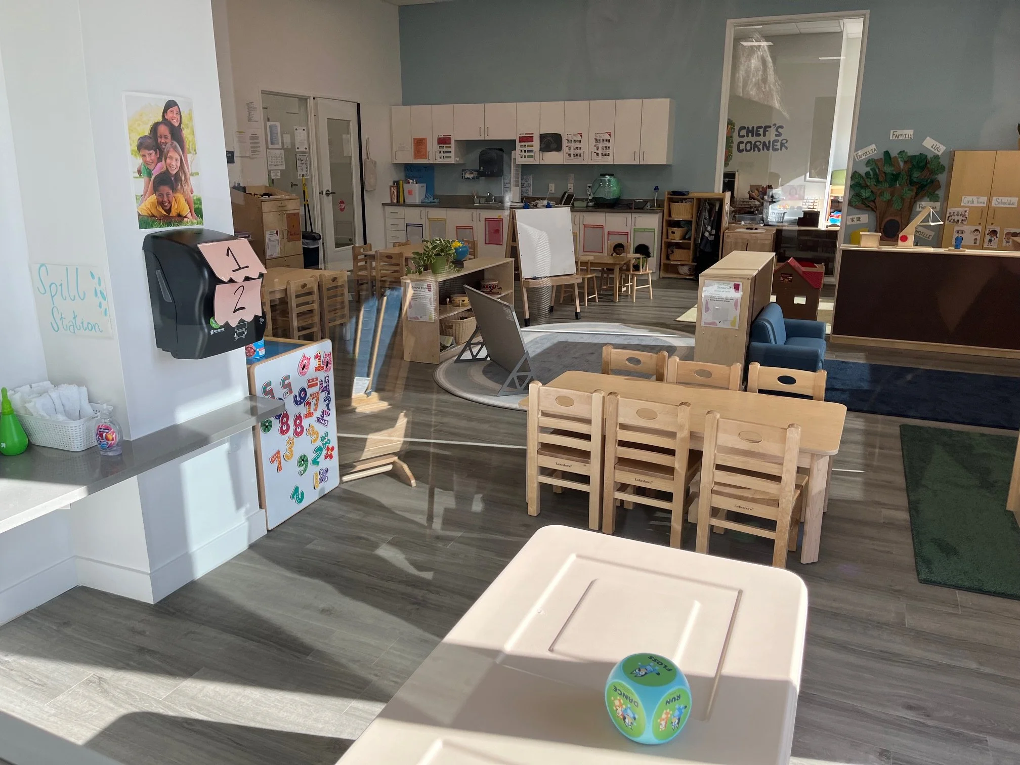 Inside a preschool classroom with various small furniture, a spill station on the wall, a table with a green toy ball, and a well-organized learning area featuring a kitchen corner, small chairs, tables, and educational materials.