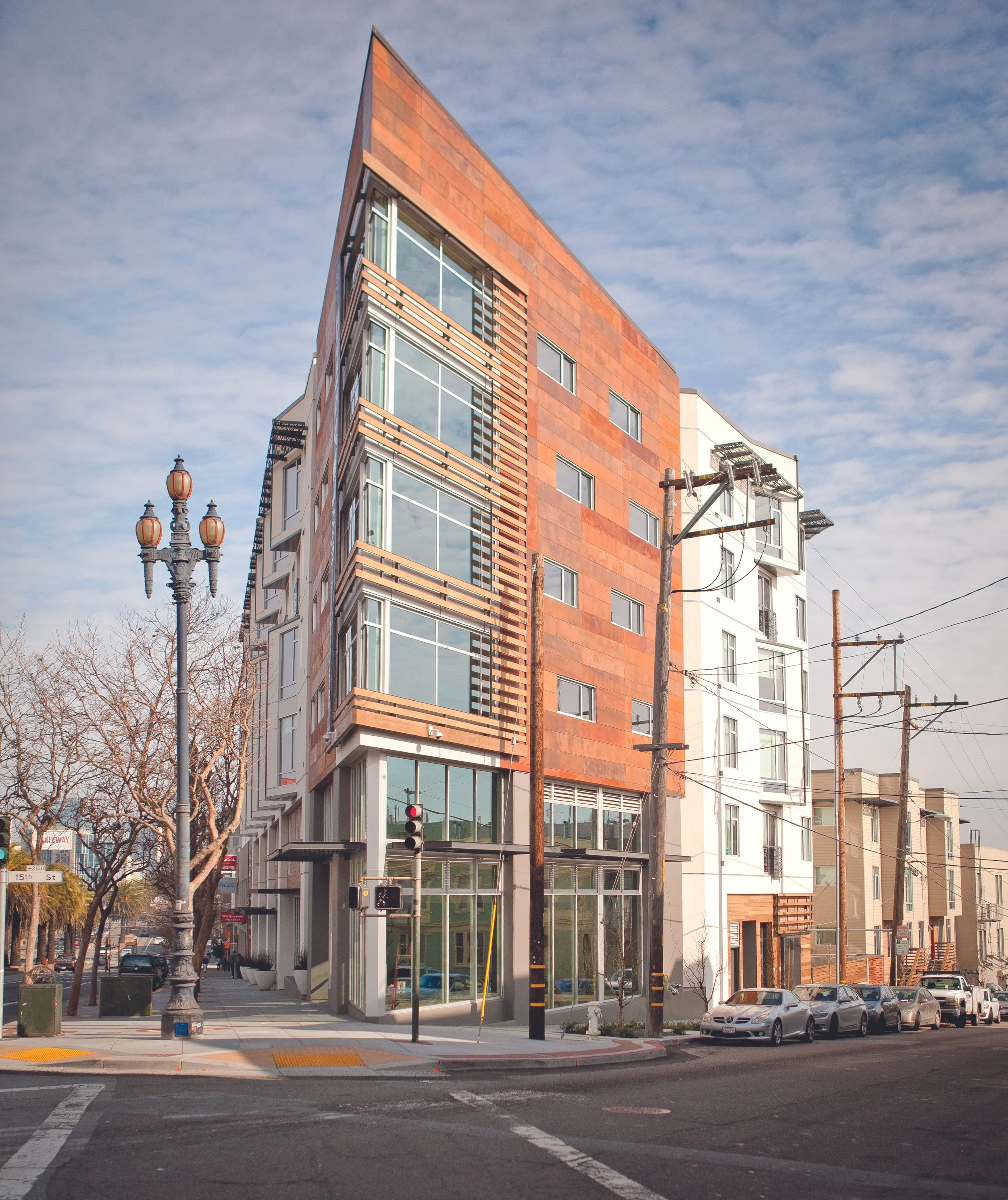 Modern multi-story building with large glass windows and a mix of reddish-brown and white exterior panels, situated at an urban street corner with parked cars and a streetlight.