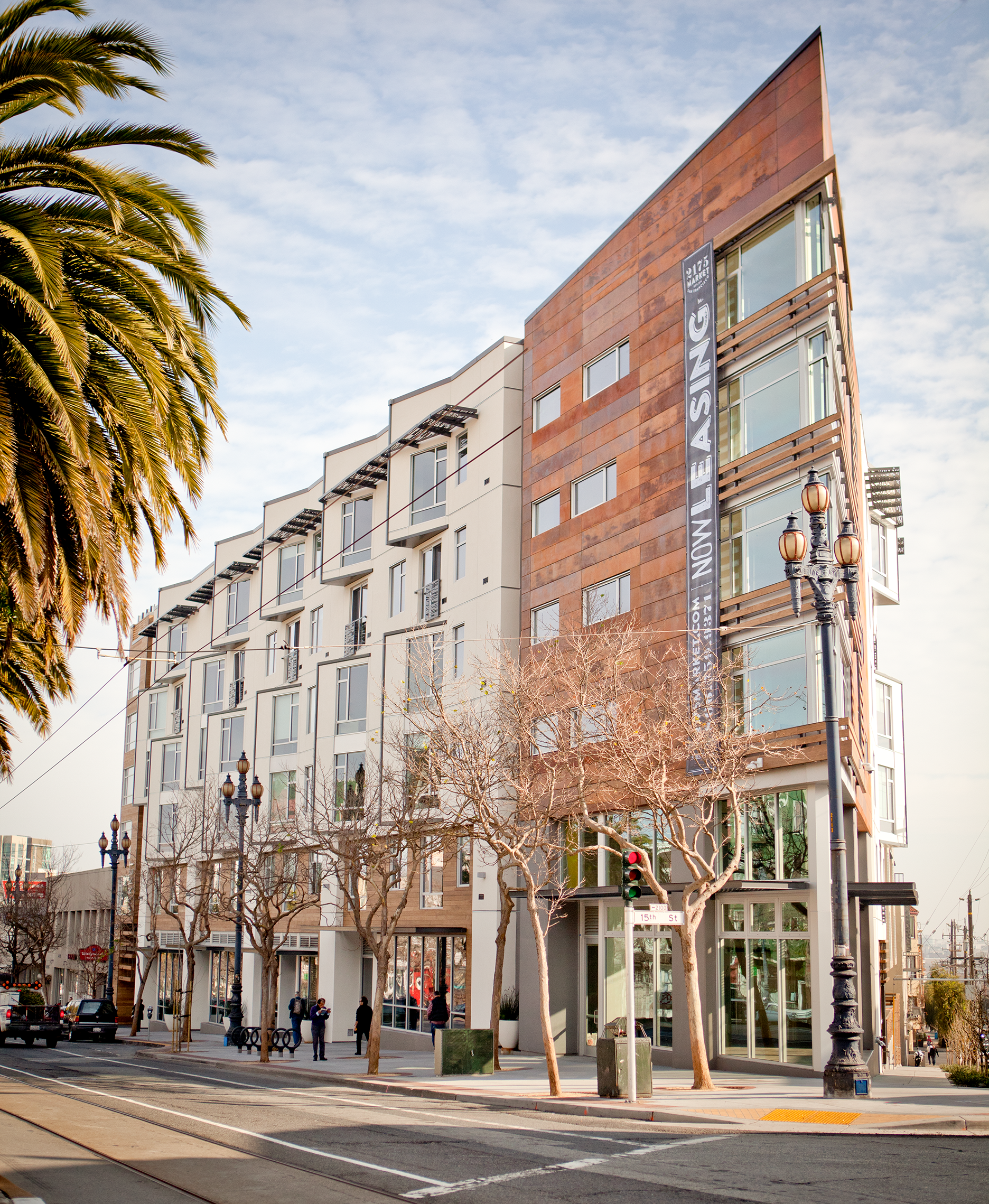 Modern multi-story apartment building on a city street with leafless trees, street lamps, and crosswalk, and a banner for the Nowleasing.com website.
