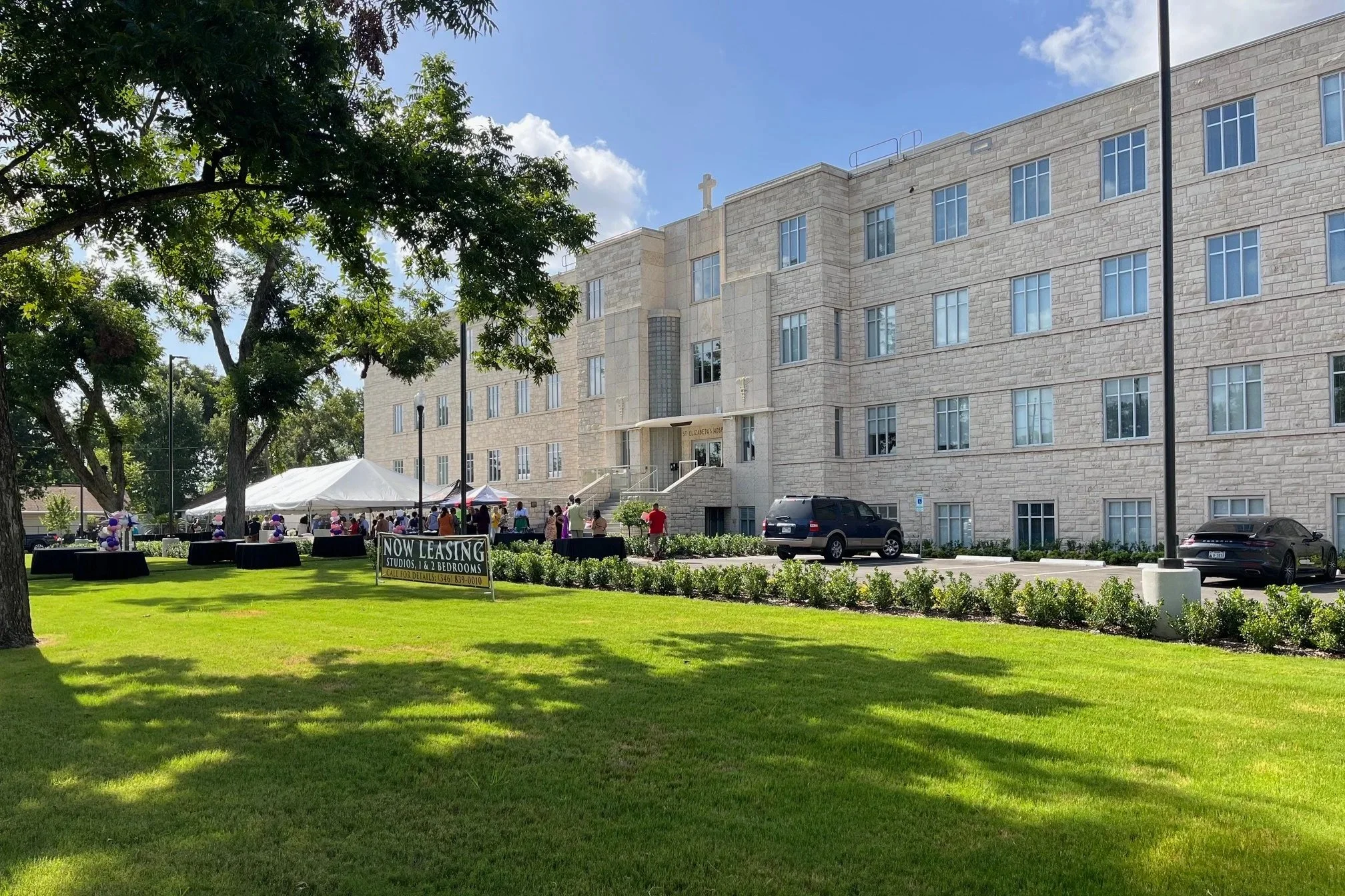 A large, beige stone church building with a cross on top and many windows, surrounded by a green lawn with trees. In front, there is a tent with people gathering, some standing near tables, and a sign that reads "Now Leasing Studios, 1 & 2 Bedrooms."