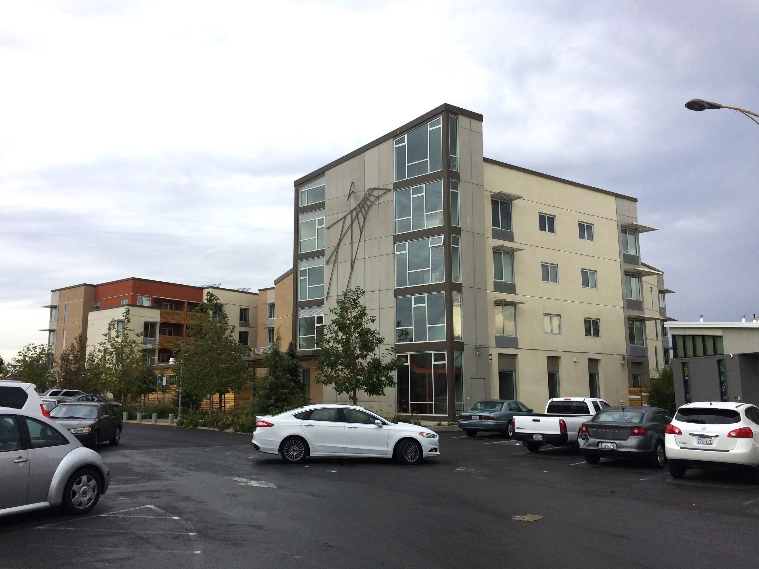 Multi-story modern apartment building with large windows and balconies, parked cars in the foreground, trees, and an overcast sky.