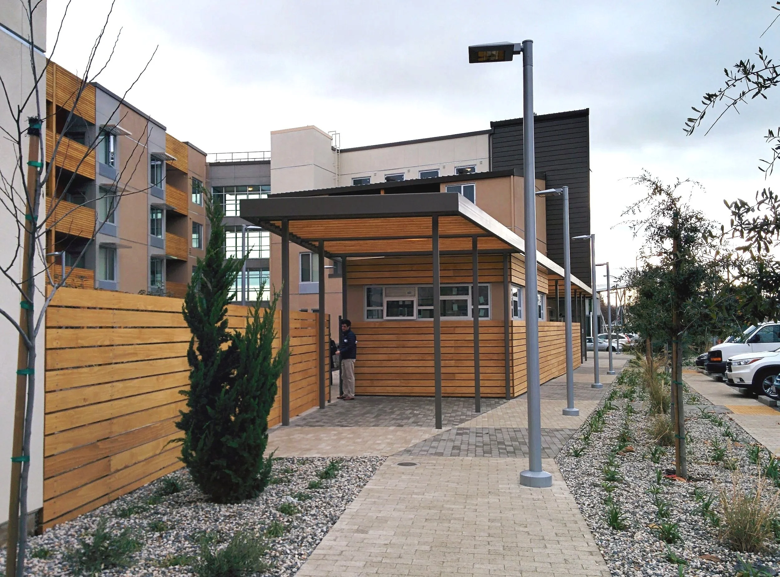 Modern building with wooden fencing, trees, street lamps, and parking area with cars.