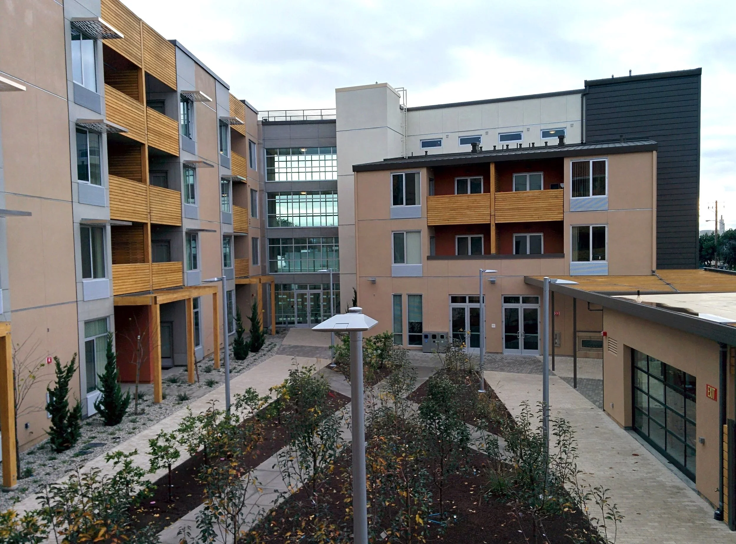 Modern apartment complex with multiple buildings, balconies, small trees, pathway, street lamps, and a landscaped courtyard.