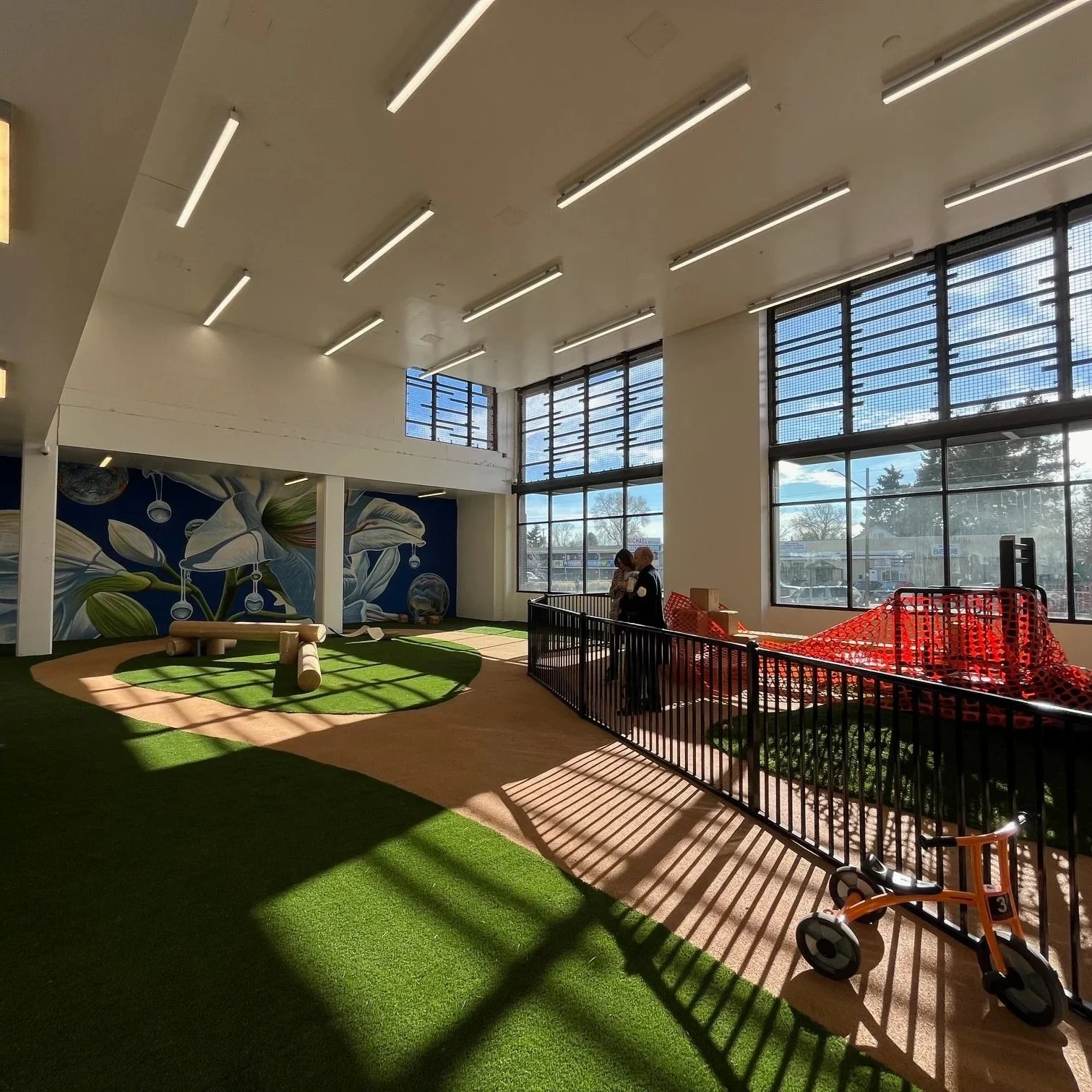 Indoor play area with artificial grass, wooden play equipment, and large windows letting in sunlight. Two people are near a red construction barrier on the right side.