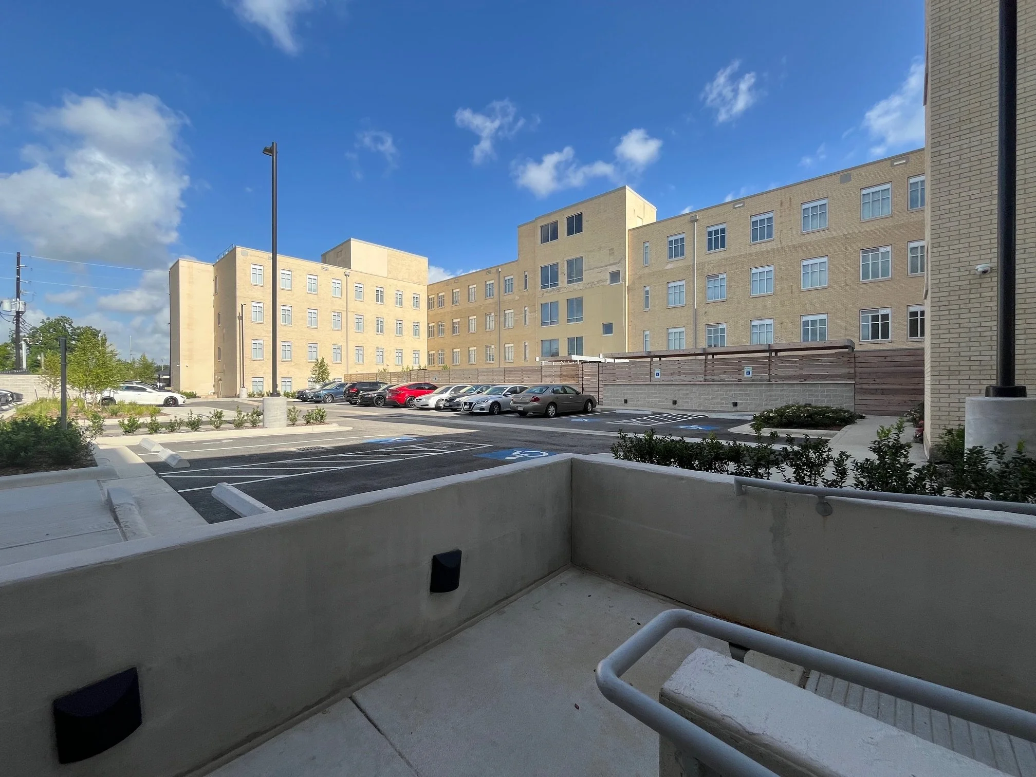 View of a parking lot with cars, a residential building in the background, blue sky with some clouds, and a small landscaped area along the sidewalk.