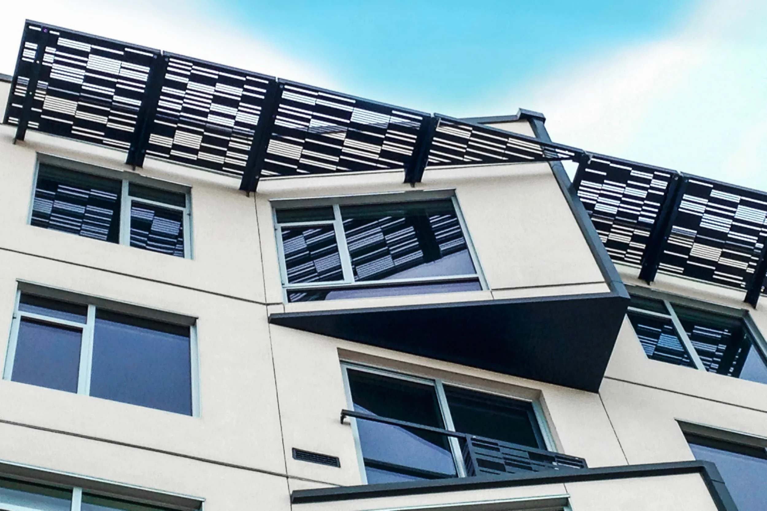 Close-up view of a modern multi-story building with large windows and black metal balcony railings, with a blue sky background.