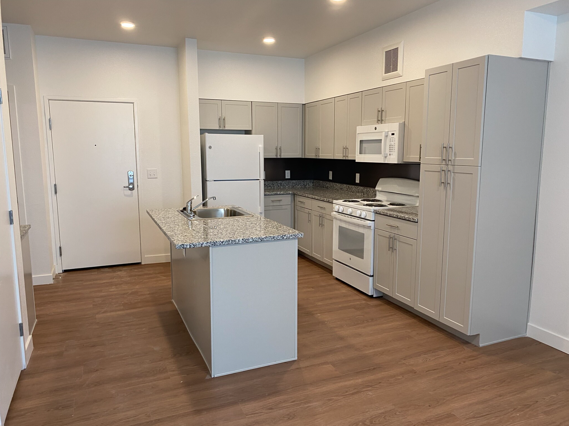 Kitchen with white cabinets, granite countertops, a white refrigerator, microwave, stove, and an island with a sink, wooden flooring, and recessed ceiling lights.