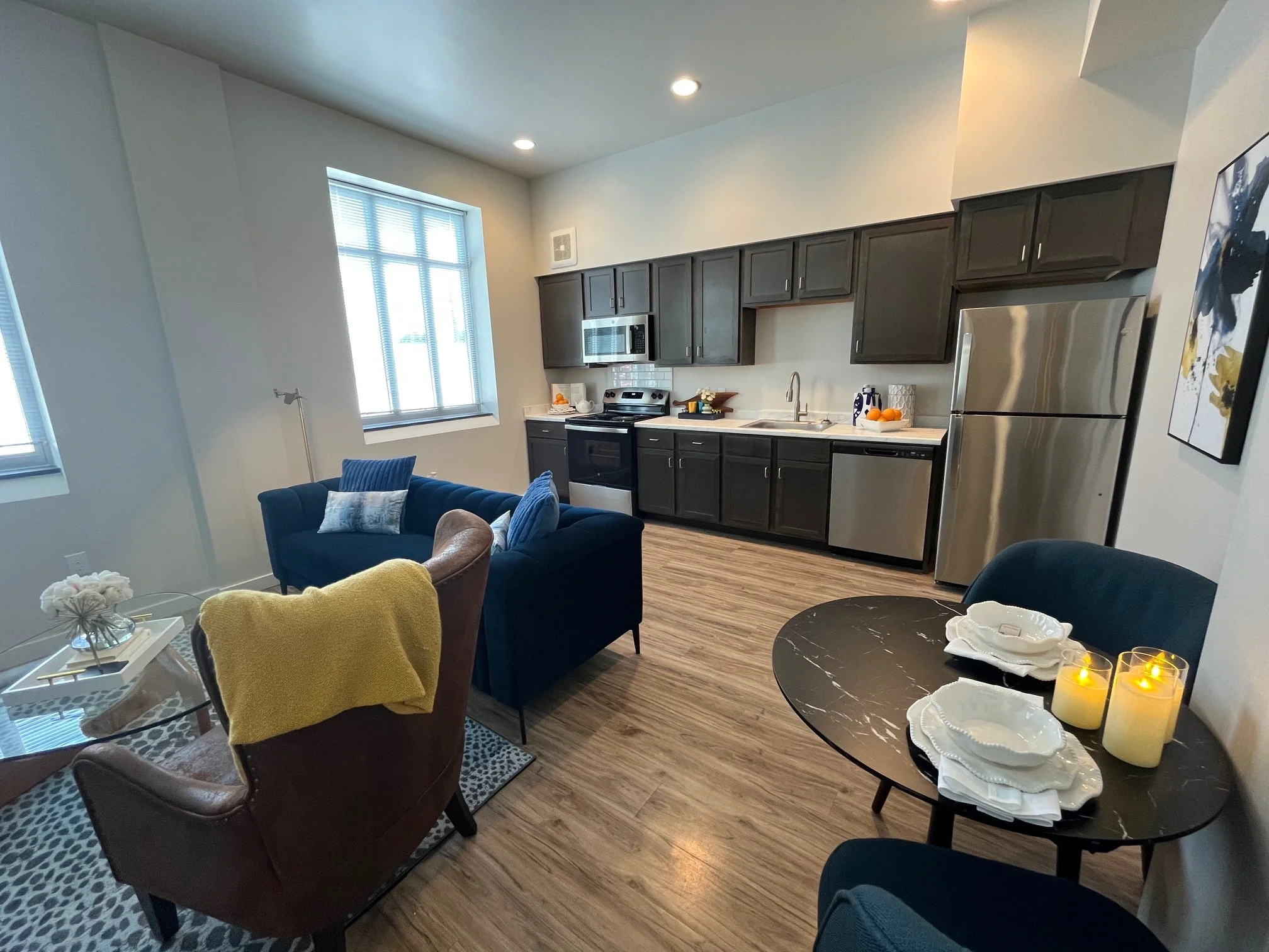 Open-plan living room and kitchen with dark cabinets, stainless steel refrigerator and appliances, a dark blue sofa with blue pillows, a brown leather chair with a yellow throw blanket, a round dining table with place settings, and candles.