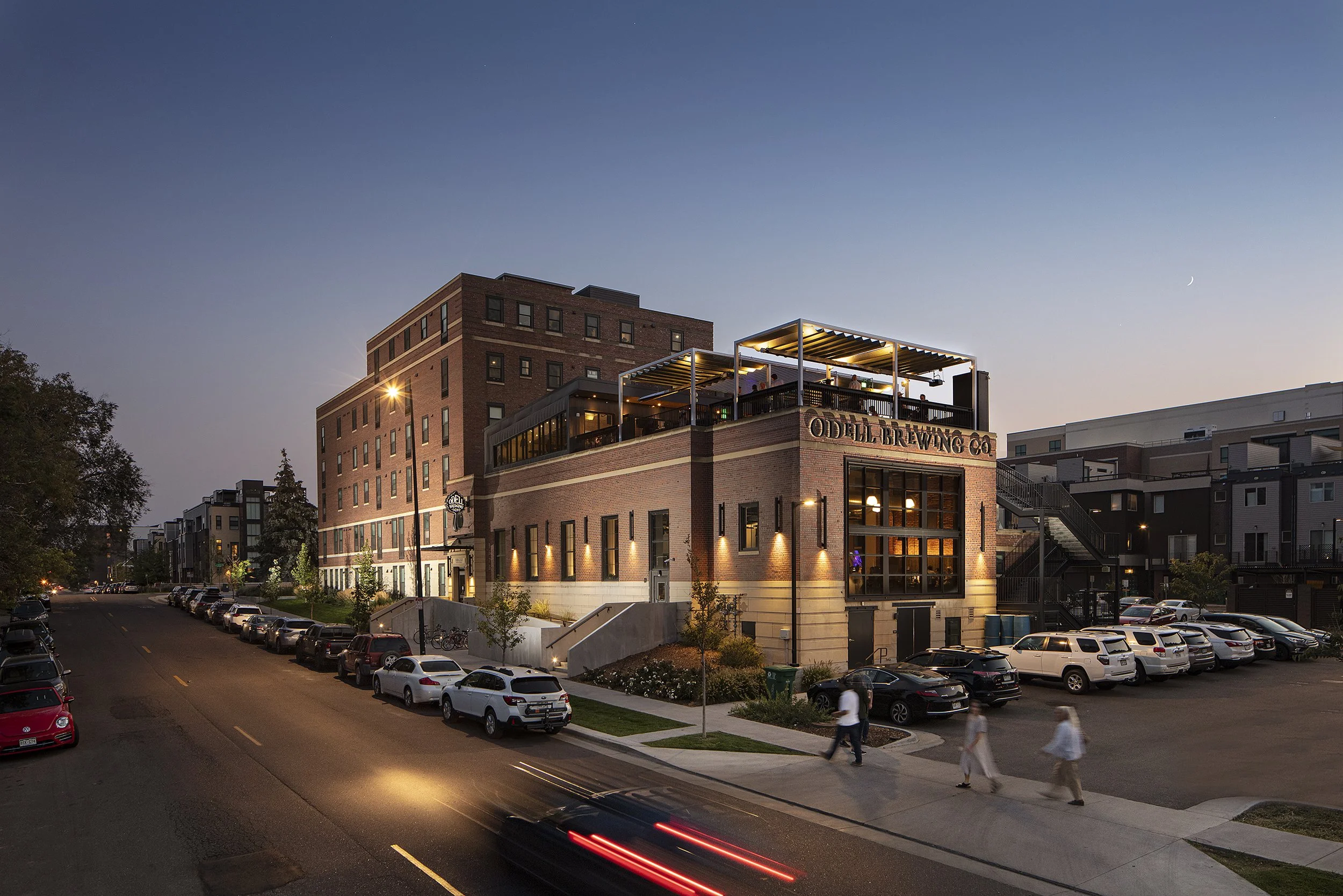 A multi-story brick building with a rooftop patio, illuminated by exterior lights, on a city street at dusk. Cars are parked along the street, and pedestrians walk on the sidewalk.