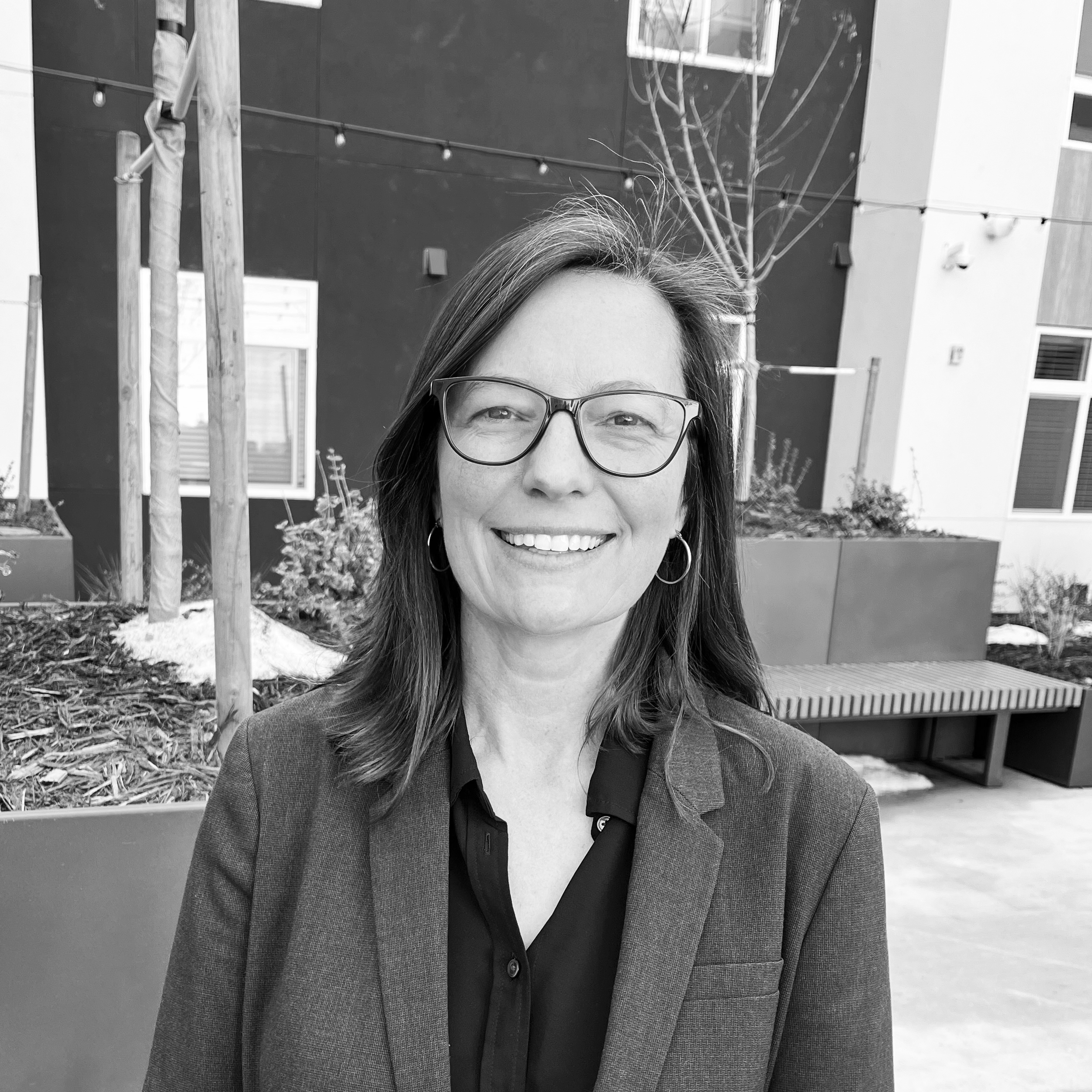 A woman with glasses, hoop earrings, and a blazer smiling outdoors in front of a modern building with planters and leafless trees.