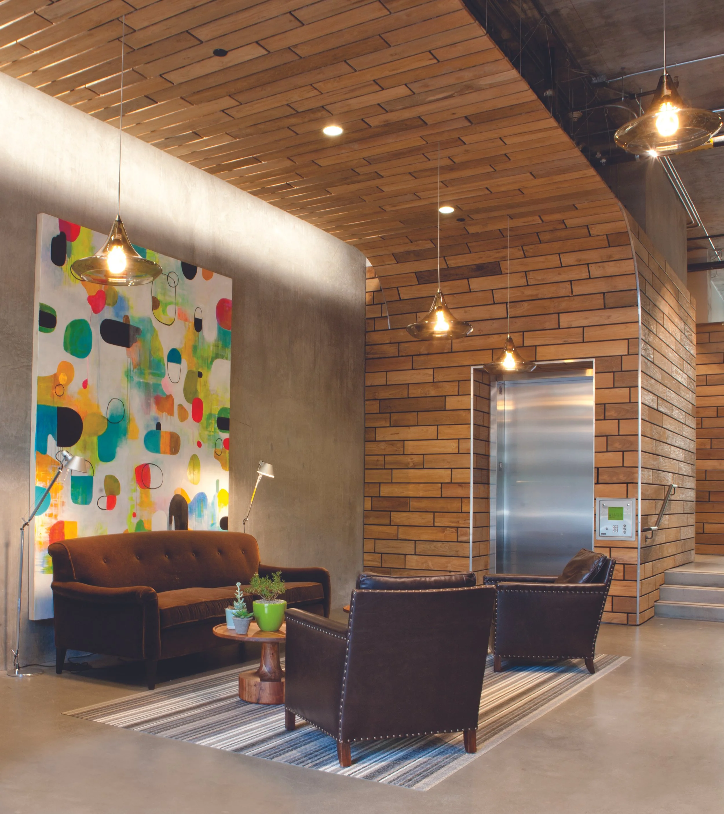 Modern lobby area featuring a colorful abstract painting, a brown velvet sofa, two black leather armchairs, a striped rug, a small wooden table with green potted plants, hanging pendant lights, a gray concrete wall, a wooden ceiling, and an elevator 