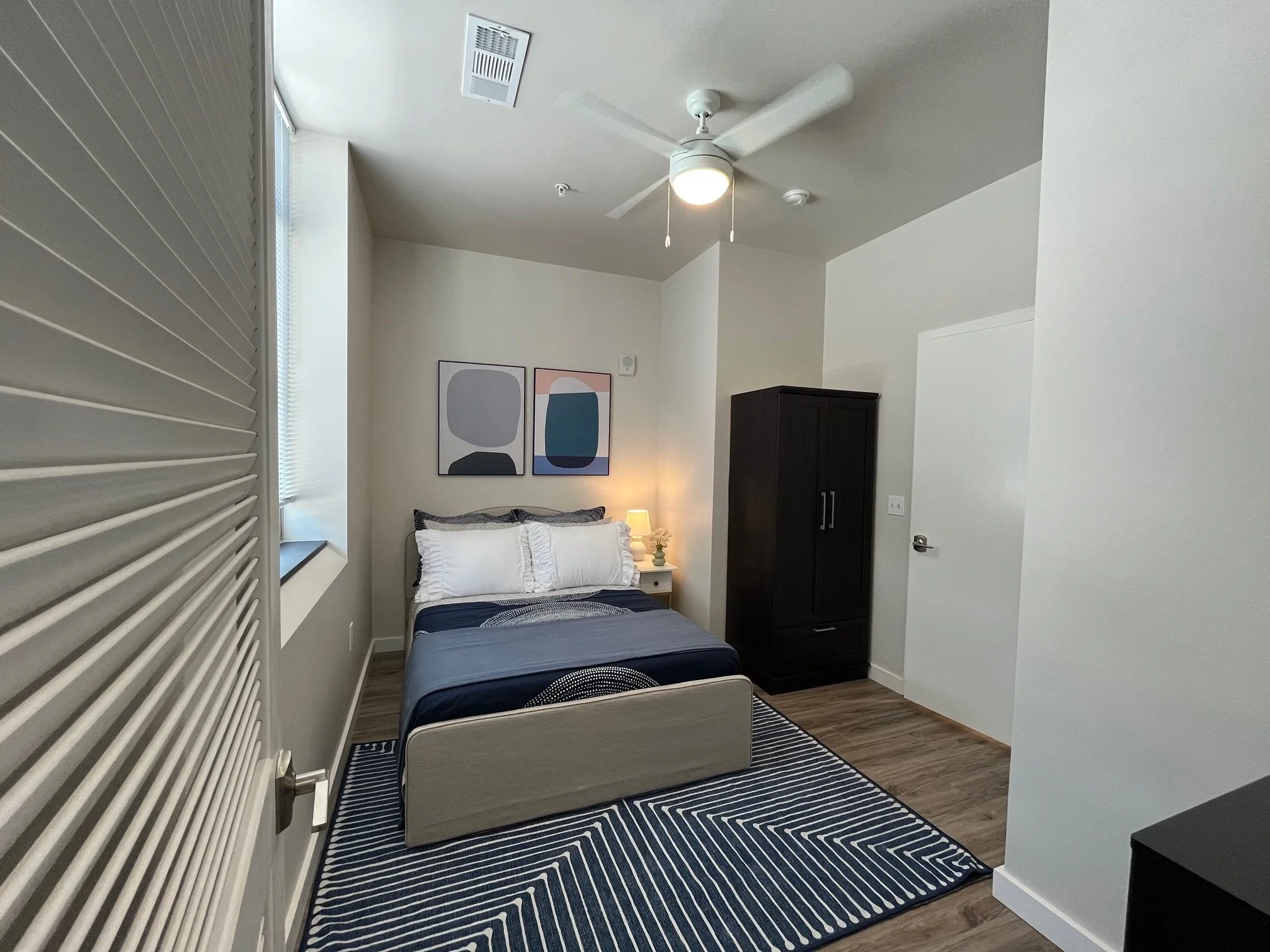 A bedroom with a bed, white pillows, navy bedding, and a geometric black-and-white rug, with abstract art on the wall, a window with blinds, a dark wardrobe, a white nightstand with a lamp, ceiling fan, and light-colored walls.