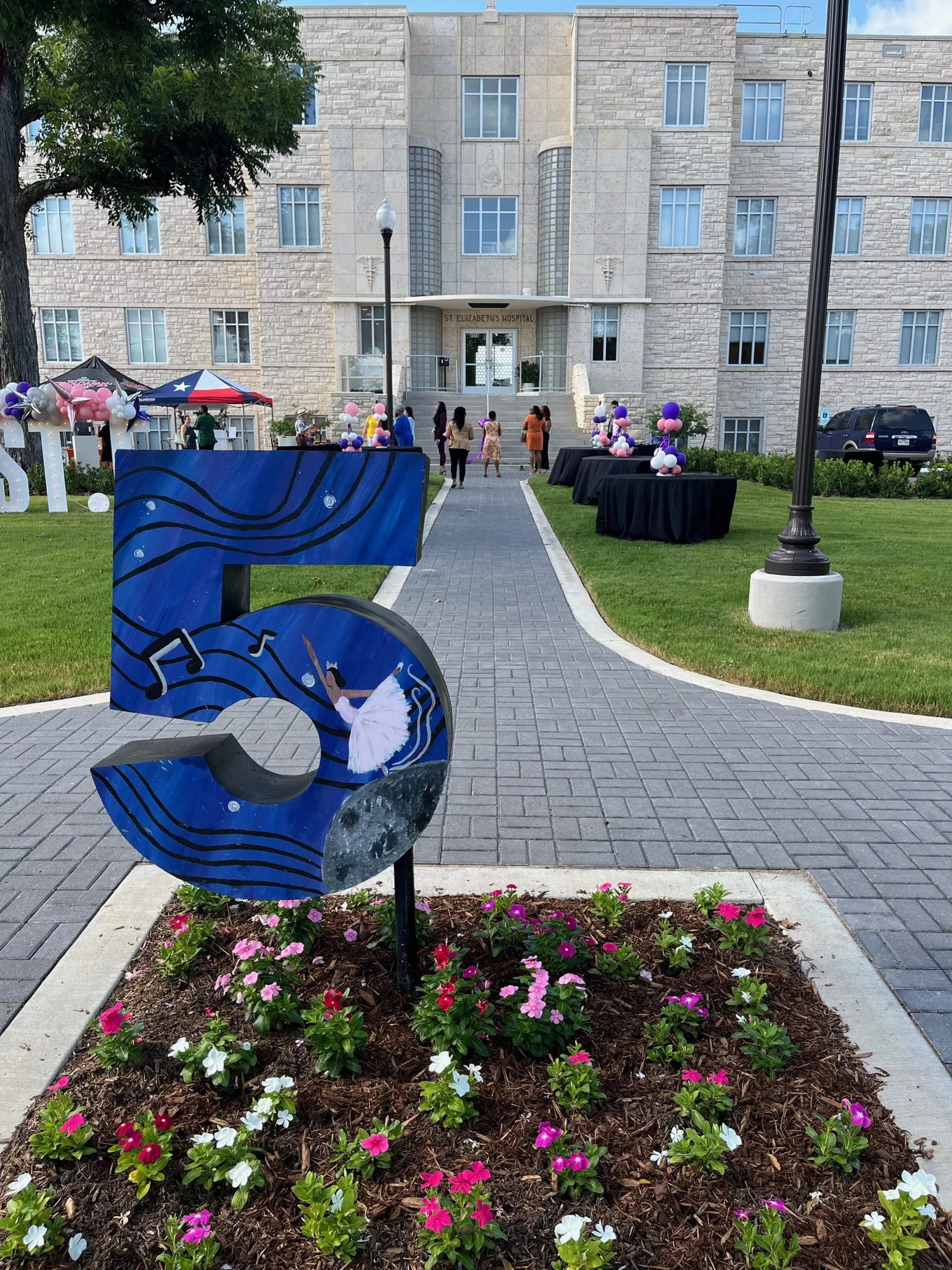Decorations and people gathered outside St. Elizabeth's Hospital, with flowers, tables with balloons, and a colorful art sign featuring a ballerina and musical notes.