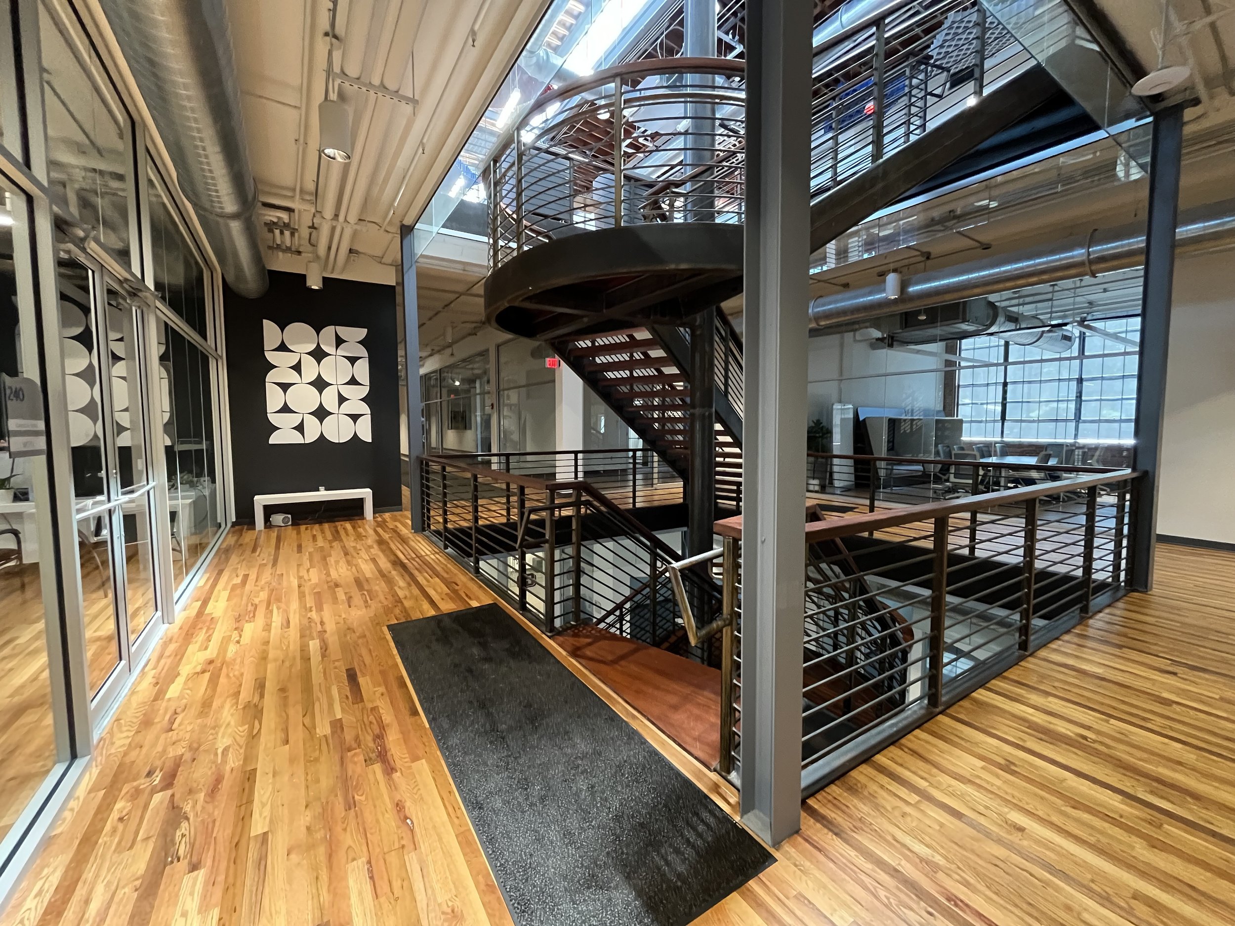 Interior view of a modern office building with wooden flooring, glass walls, a black wall with geometric white patterns, a curved staircase with wooden steps and metal railings, and large windows allowing natural light.