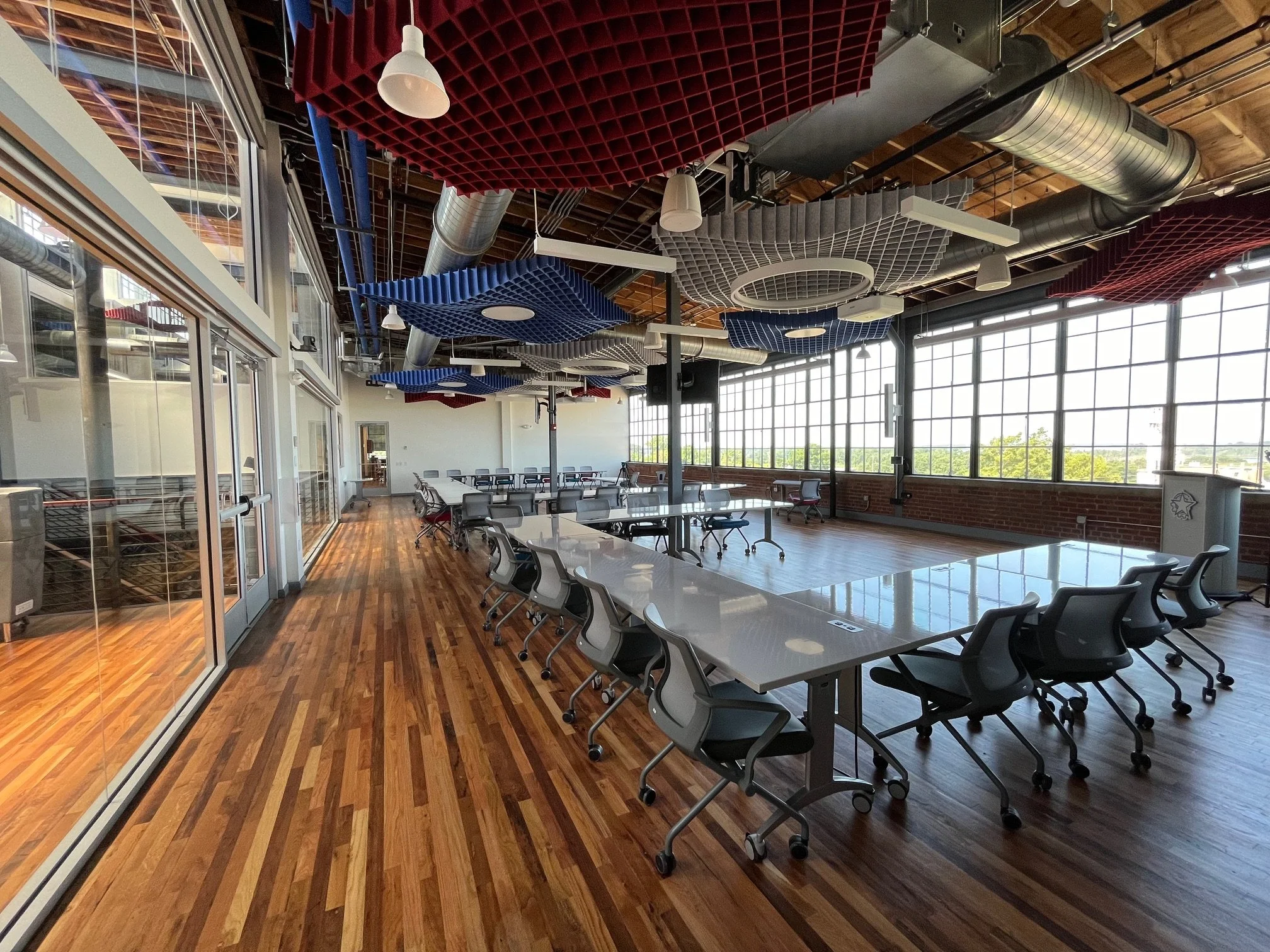 A spacious conference room with large windows, white tables, black office chairs, and colorful acoustic ceiling panels in red, blue, and gray, with exposed ductwork and wooden floors.
