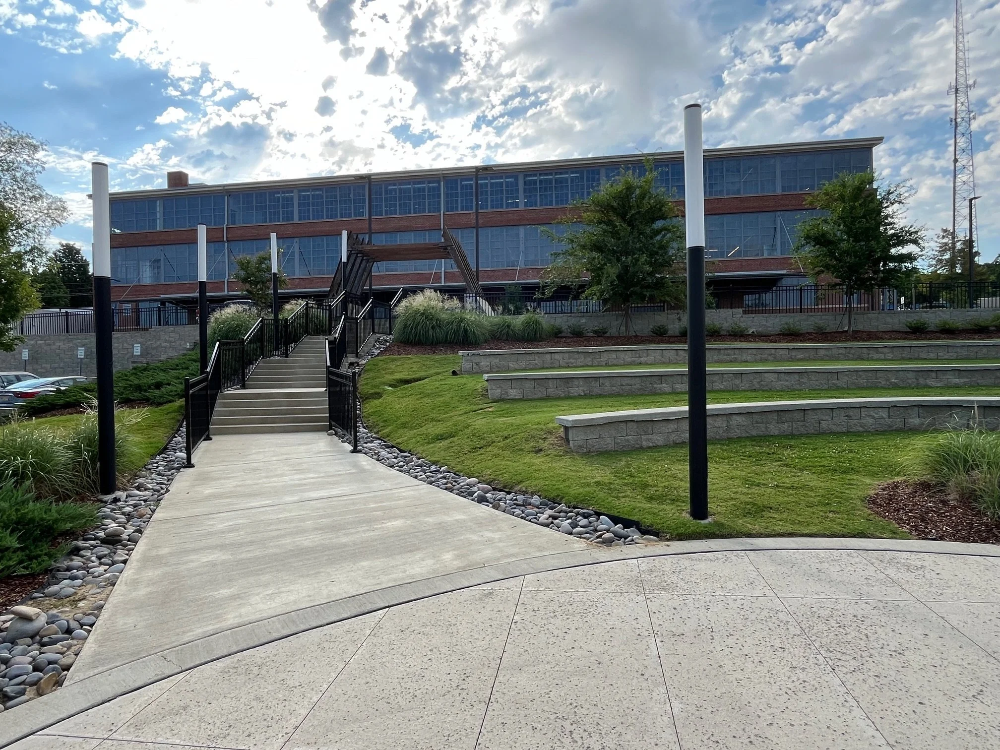 A concrete pathway with metal railings and stone edging leading up a small hill toward a modern building with large windows, surrounded by trees and landscaped grass.