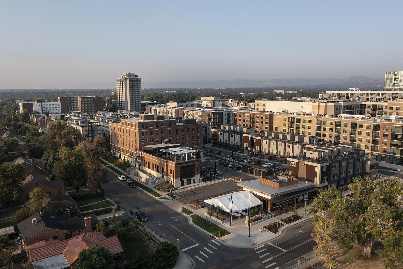 Aerial view of a cityscape showing modern mid-rise buildings, a few older houses with red roofs, trees, and streets with parked and moving cars.