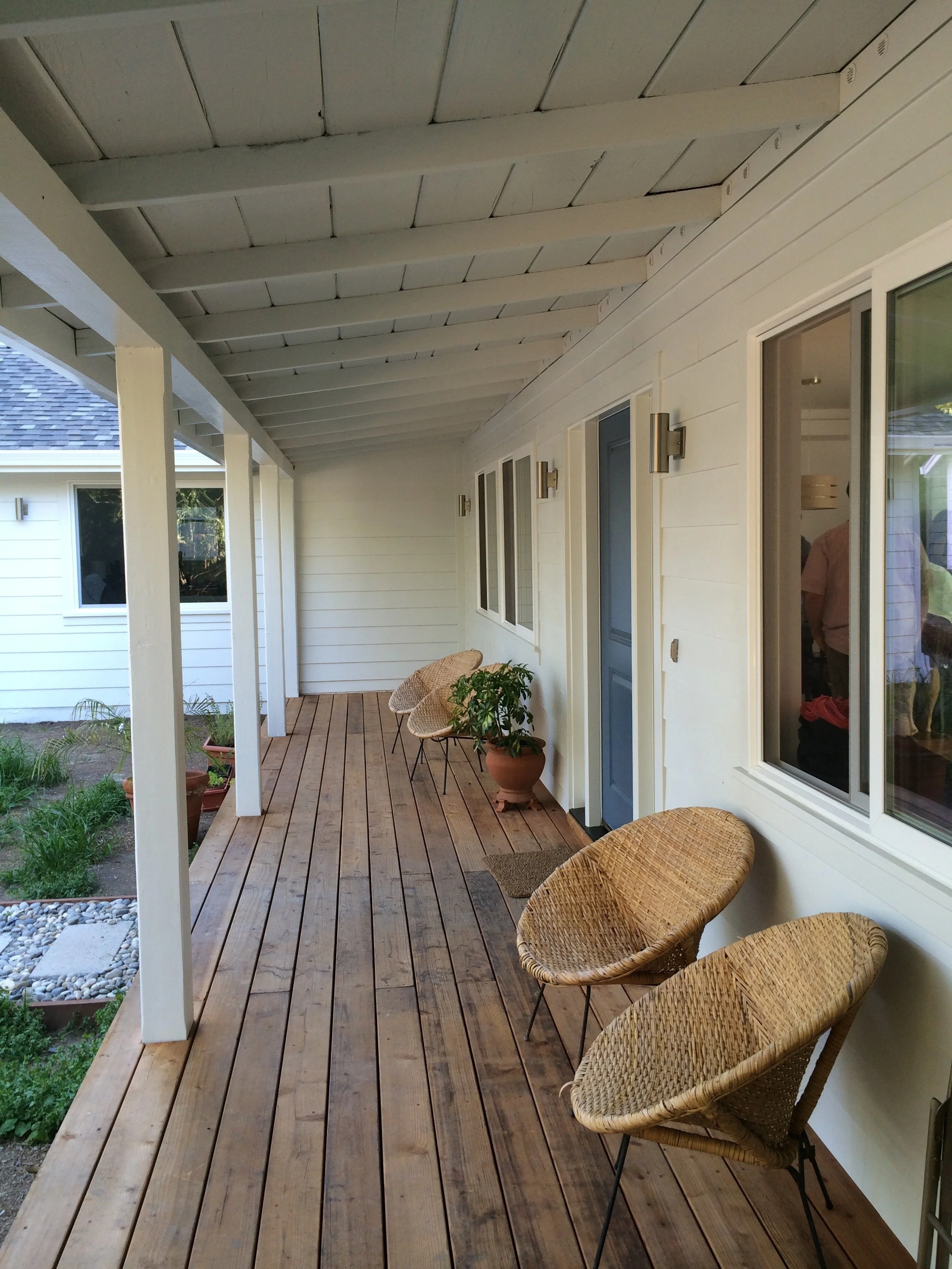 A spacious porch with a wooden floor, white exterior walls, large windows, and a door painted blue. There are wicker chairs and potted plants along the porch, with a view of a garden area.