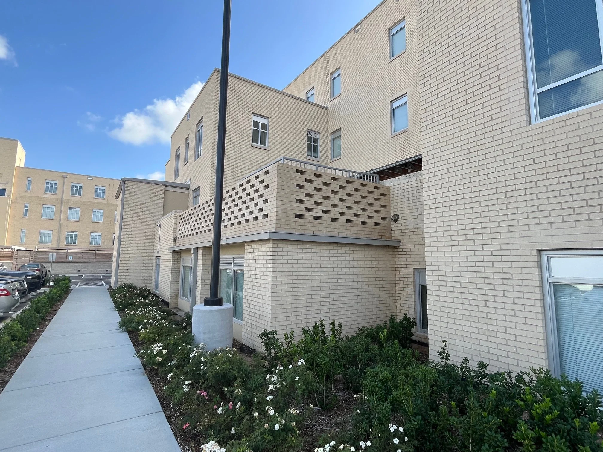 Exterior of a beige brick apartment building with several windows, a sidewalk, parked cars, and landscaped bushes with flowers under a partly cloudy sky.