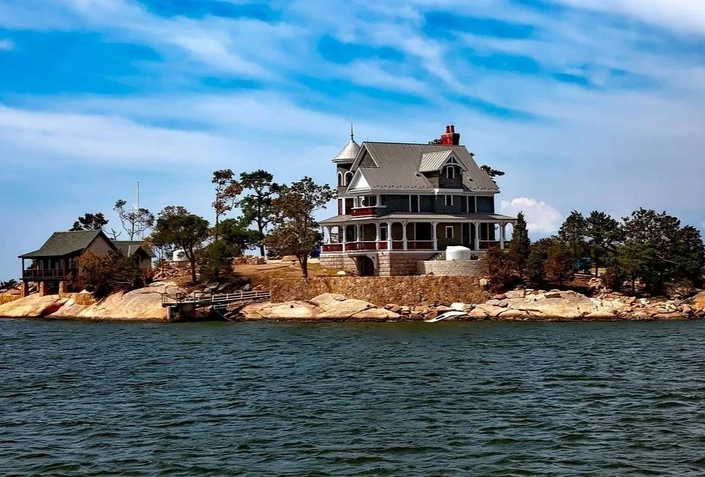 A large, Victorian-style house on a rocky shoreline by a body of water, with a smaller house and trees nearby under a partly cloudy sky.
