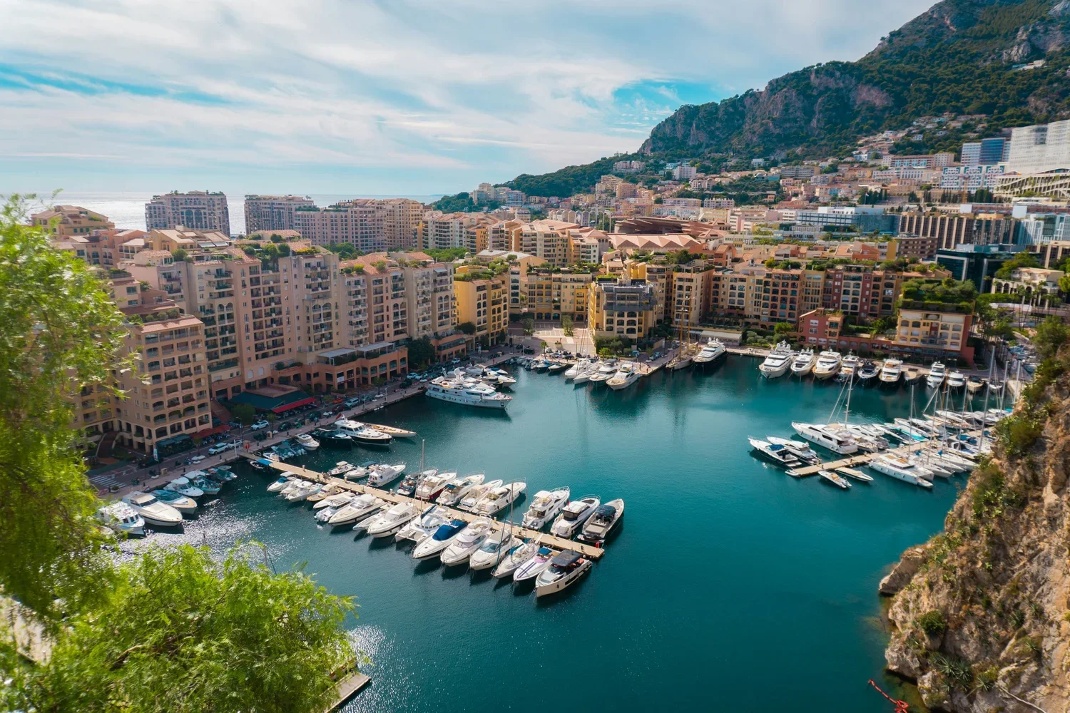 A harbor with numerous yachts and boats docked in front of pastel-colored apartment buildings, with a hillside covered in greenery and buildings in the background, under a partly cloudy sky.