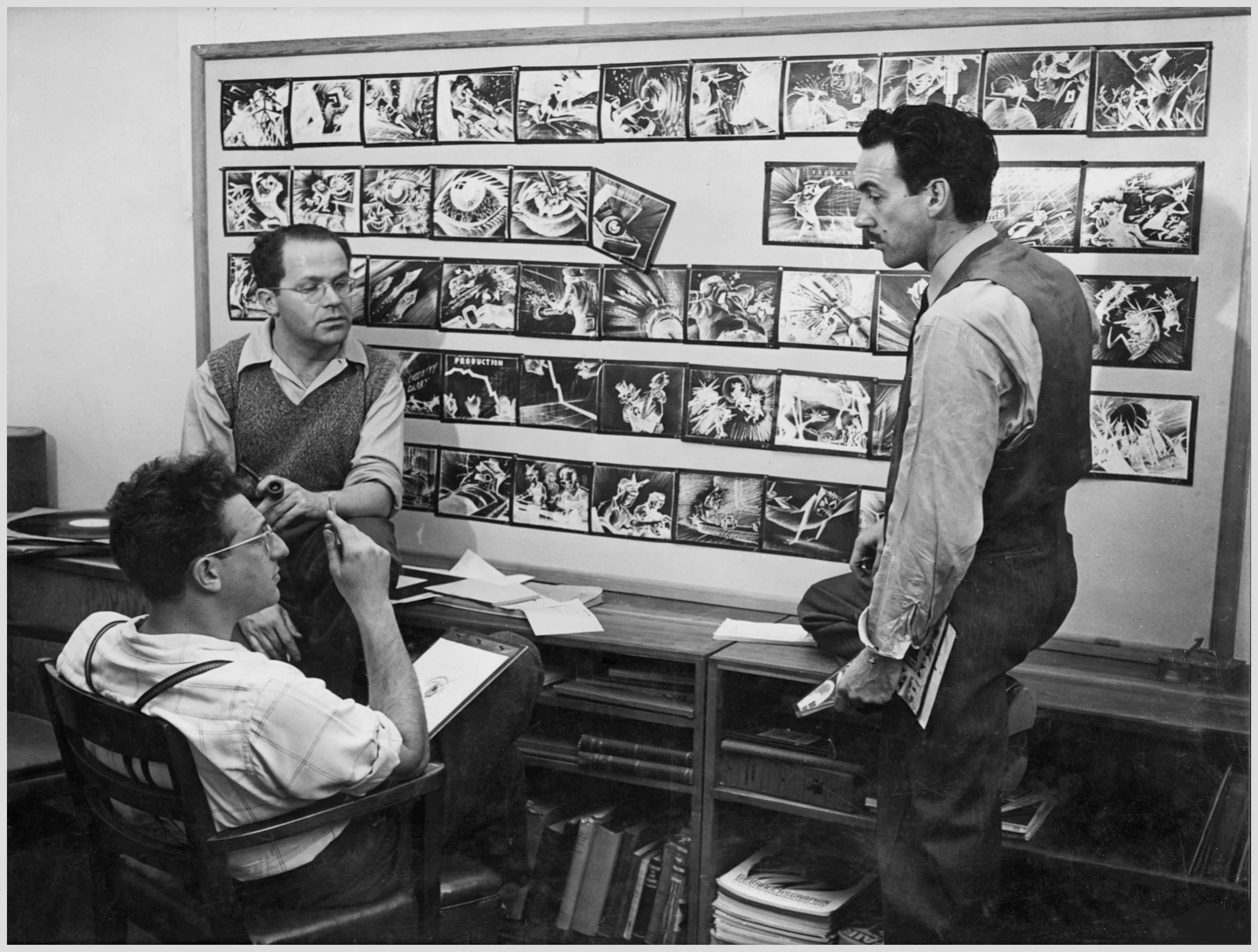 Black and white photo of three men, Zachary Schwartz, David Hilberman, and Stephen Bosustow. in a studio space discussing storyboards displayed on the wall.