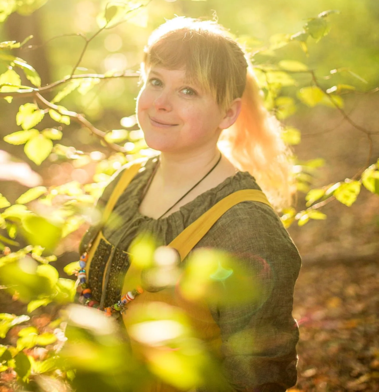 Rachel Gitlevich, a woman with short hair and a ponytail, wearing a dark brown top and a beaded necklace, stands among green leaves with sunlight shining through in a forest setting.