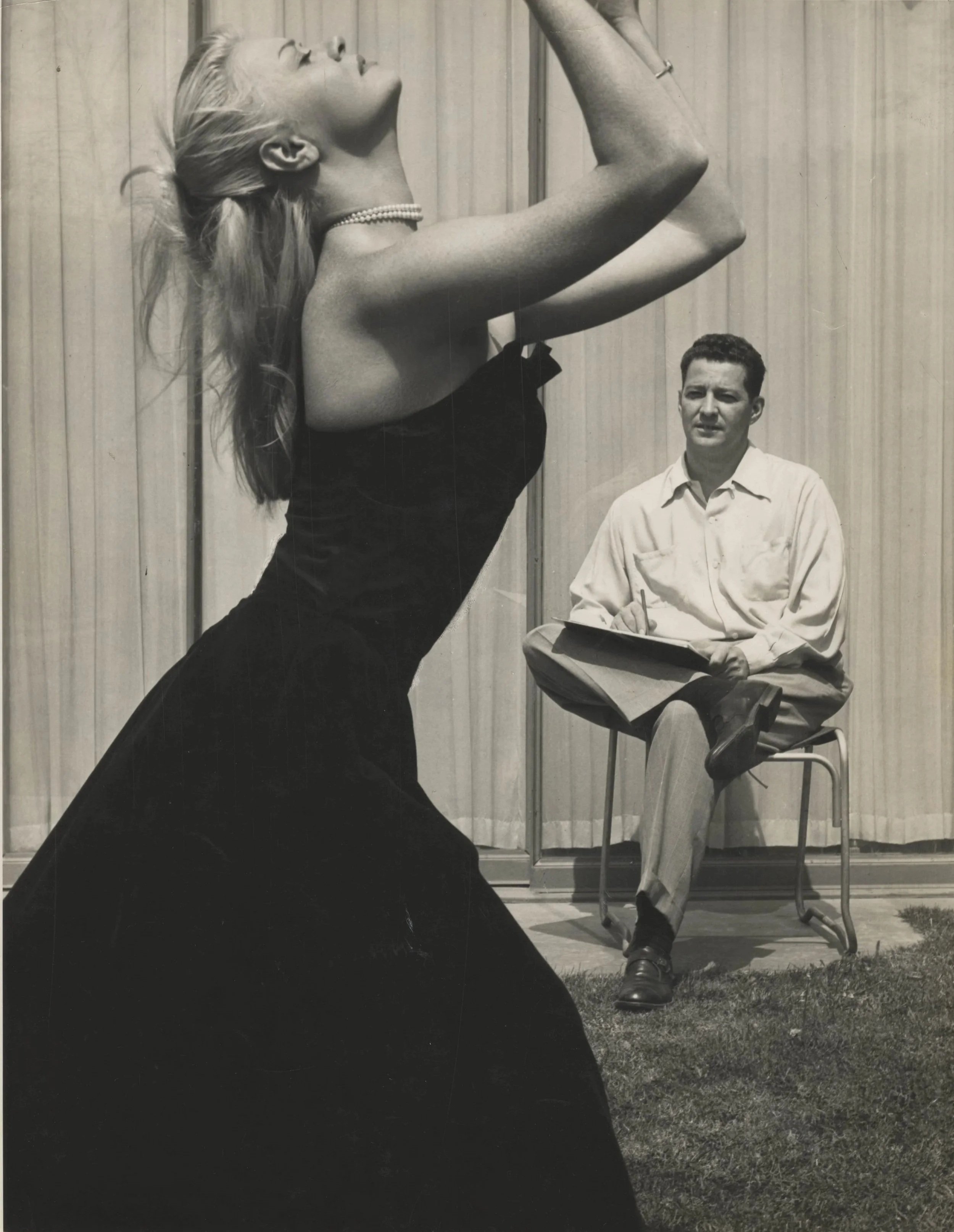 A black and white photograph of Olga Lunick, a woman with long hair, wearing a strapless dress and a pearl necklace, posing in front of a wall with vertical siding. John Hubley is sitting on a chair in the background with a notepad.