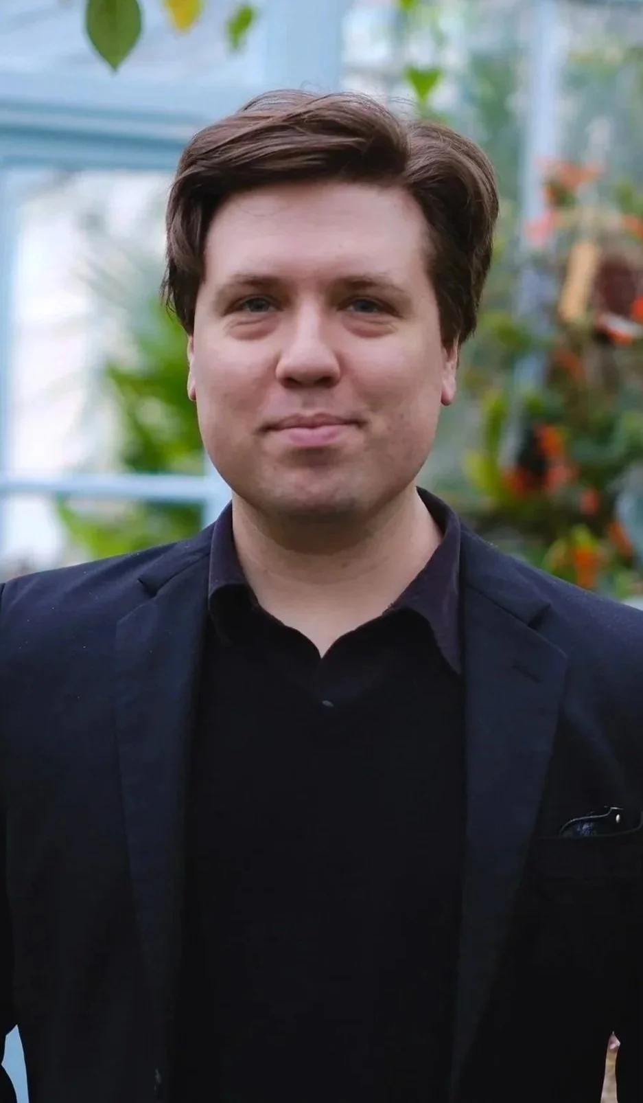 Portrait of a young man with brown hair, wearing a black blazer and shirt, standing in a greenhouse with blurred green plants in the background.