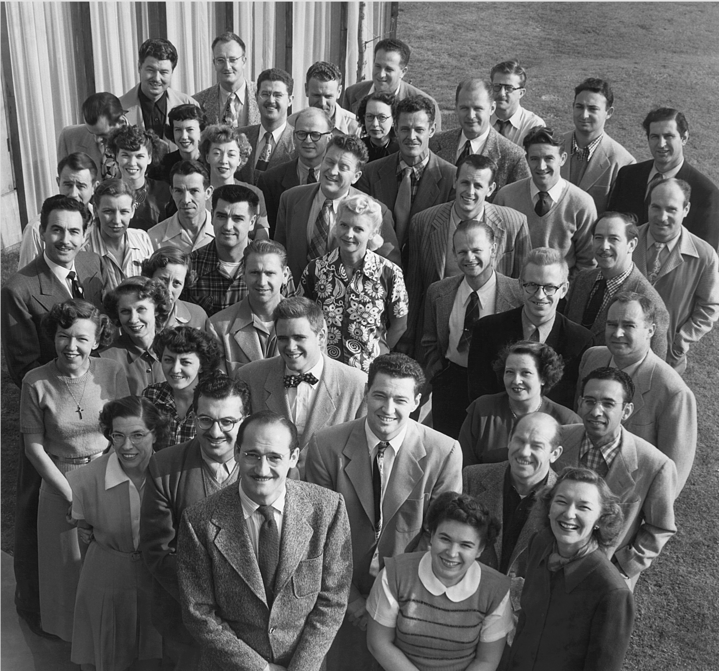 Black and white photo of a large group of men and women from the UPA animation team, dressed in vintage clothing, standing outdoors in front of a building and grass, smiling at the camera.