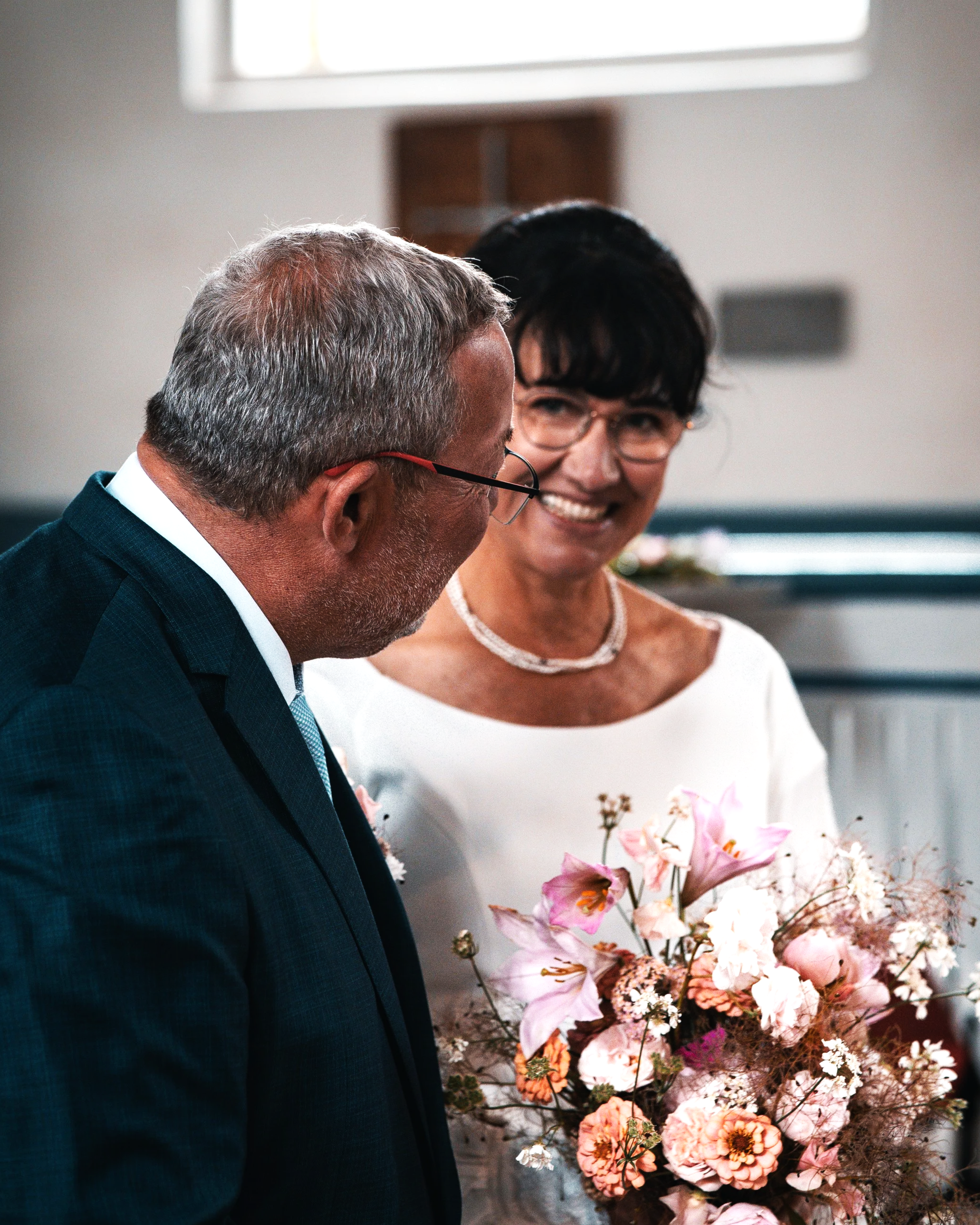 Ein älteres Paar bei einer Hochzeit, der Mann trägt einen Anzug, die Frau hält einen Blumenstrauß und trägt eine weiße Kleidung.