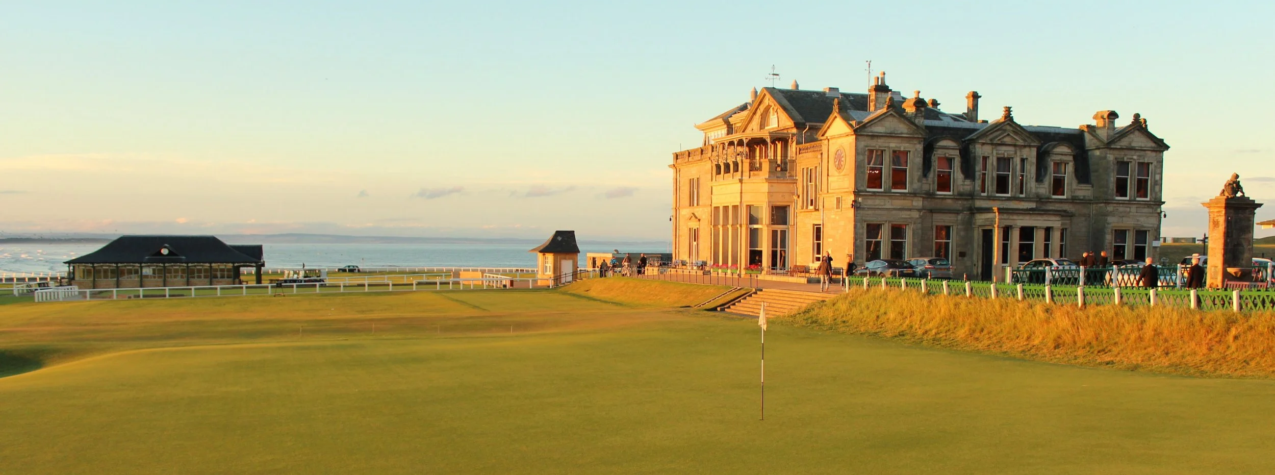 A large historic building with intricate stone architecture sits on a hill near the coastline, with cars and people around it during golden hour.