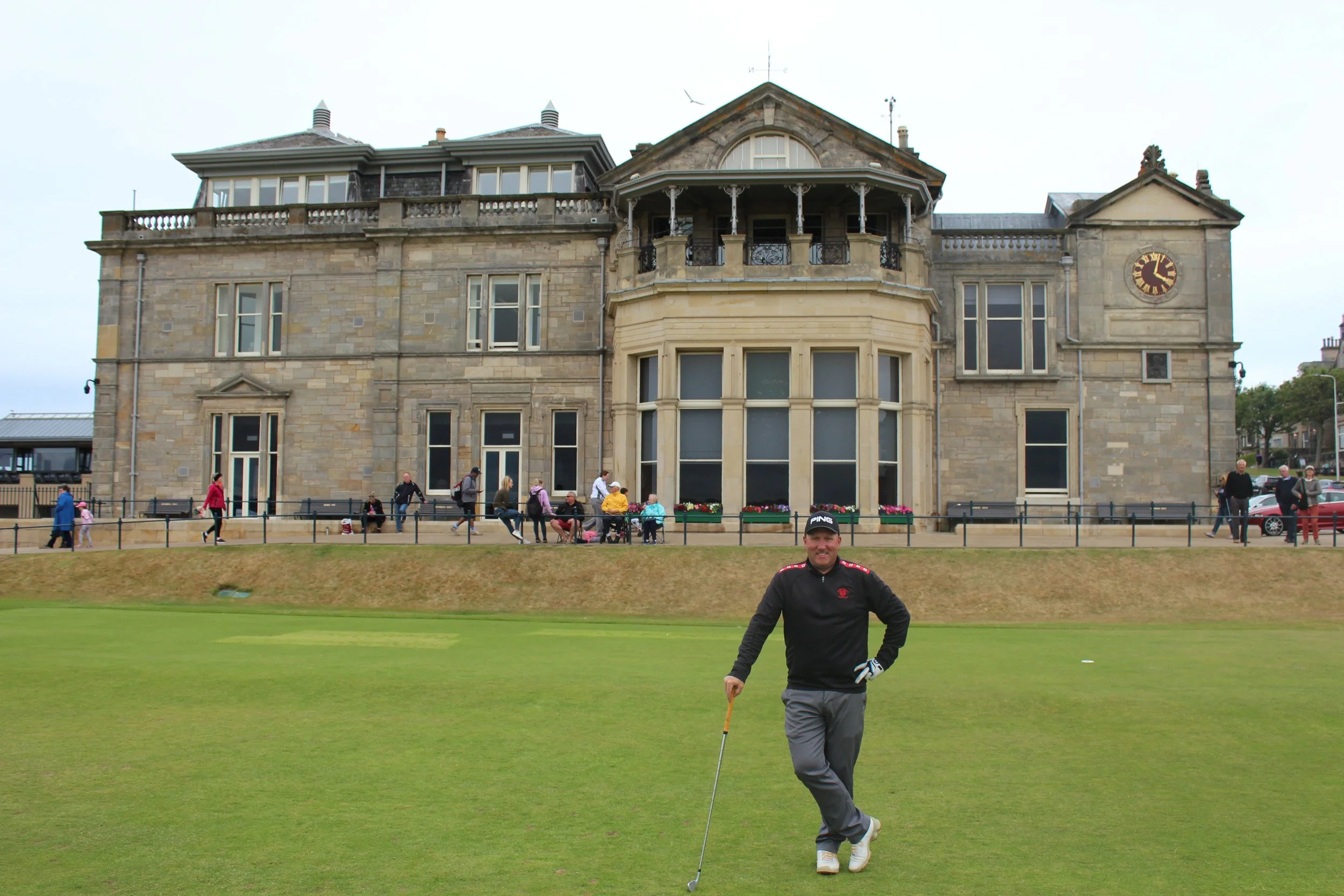 A man in a black jacket and gray pants standing on a golf course holding a golf club, with a large historic building in the background, and people walking and sitting outside.