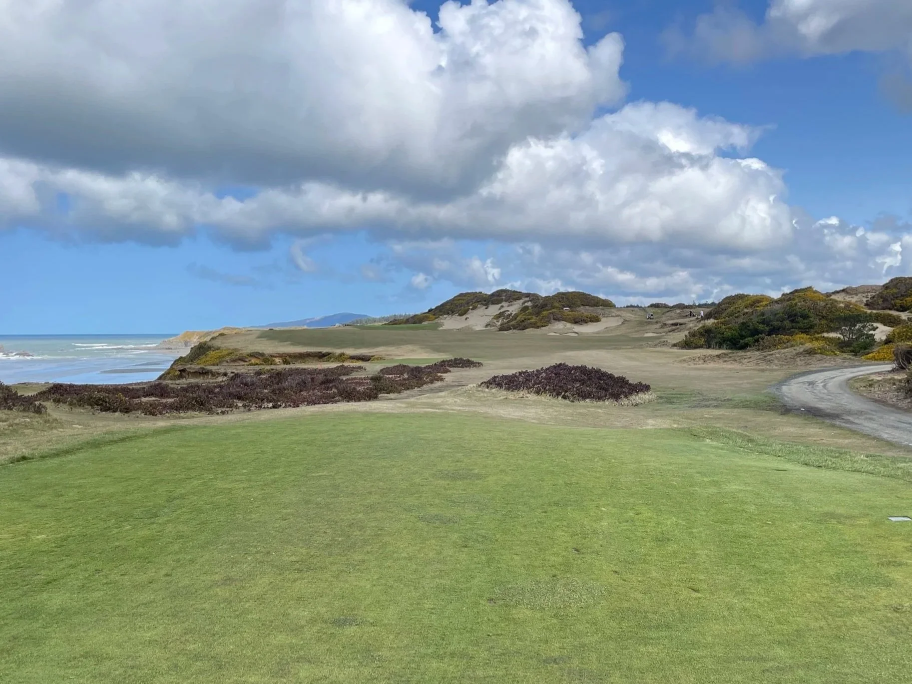 A golf course with green grass, sandy dunes, and bushes along the coastline with ocean waves visible in the background under a partly cloudy sky.