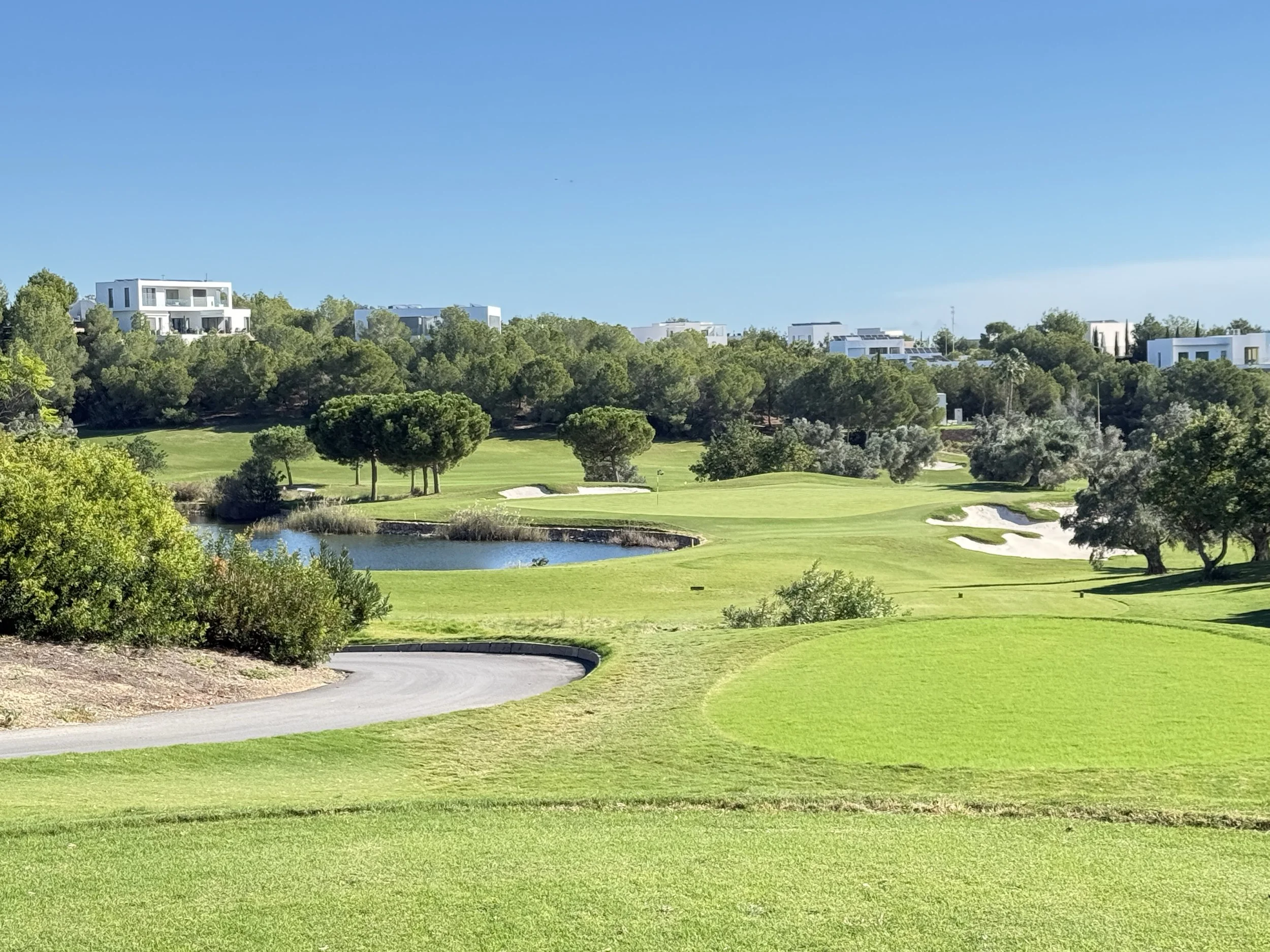 A scenic view of a golf course with green fairways, sand bunkers, a water hazard, and trees under a clear blue sky, with modern white residential buildings in the background.