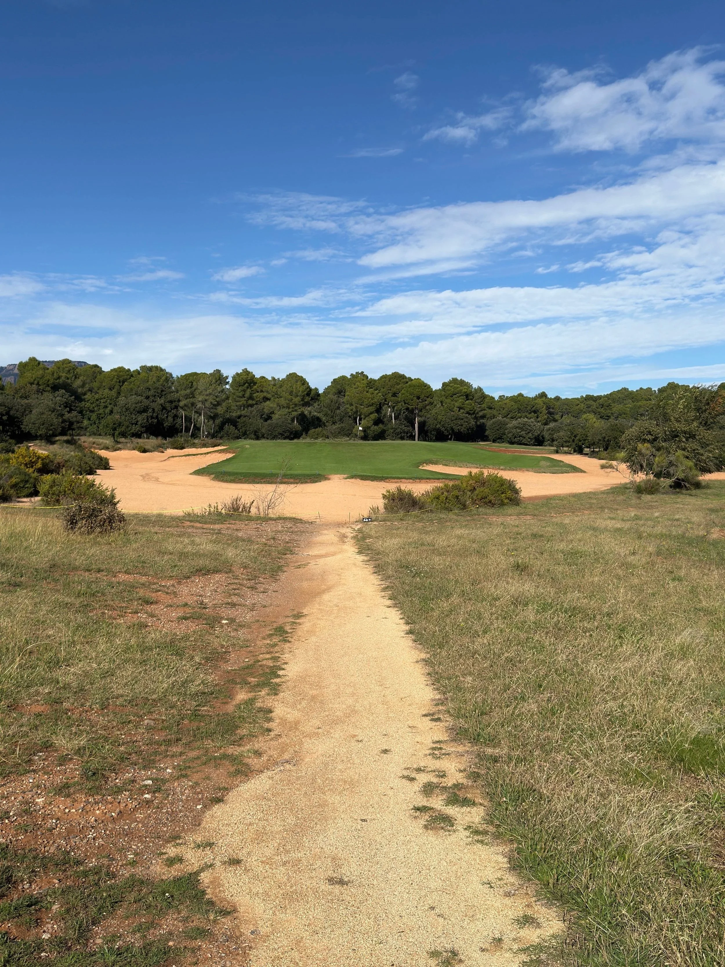 A dirt path leading to a golf course green, with sand traps on either side and trees in the background, under a partly cloudy blue sky.