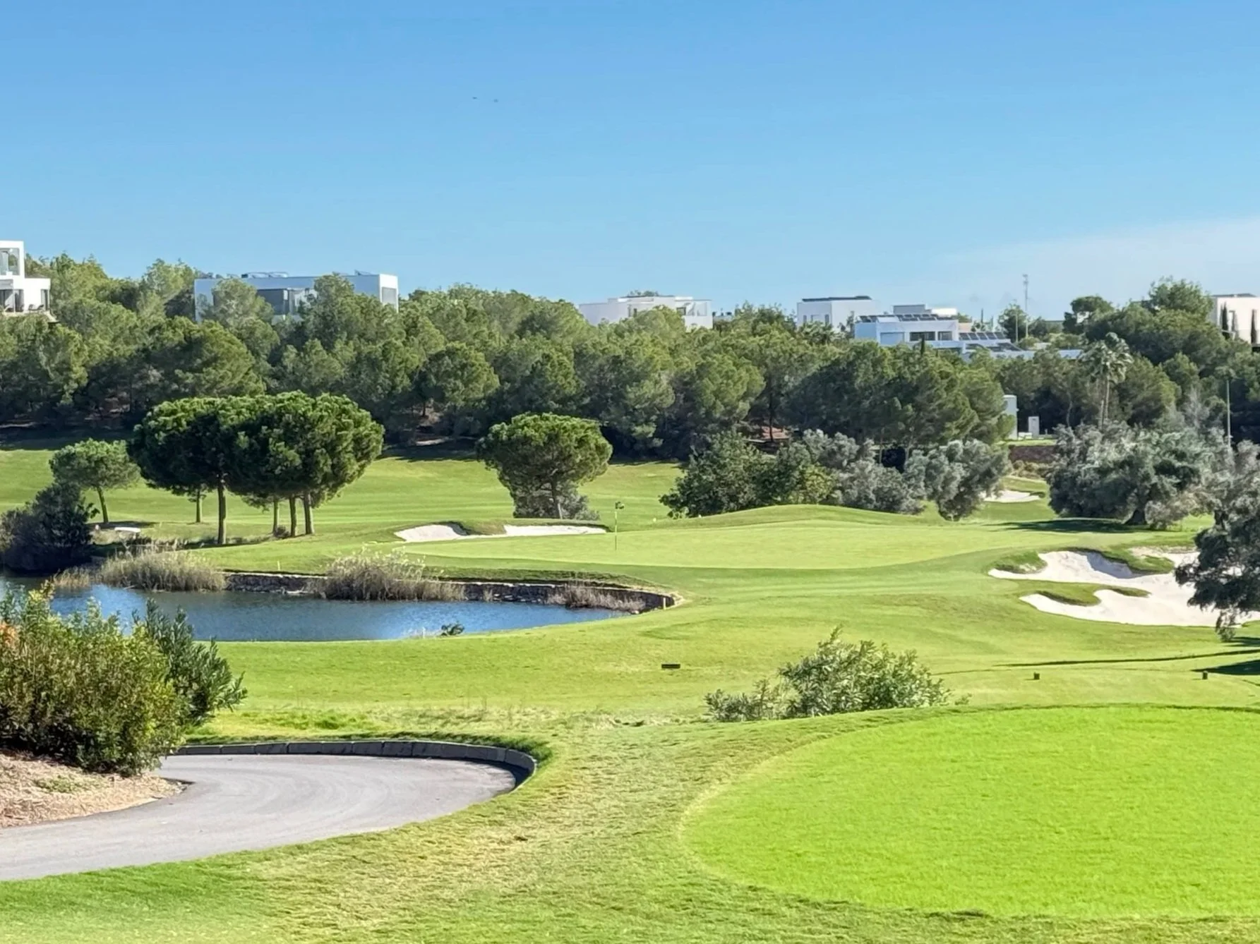 A golf course with green grass, sand bunkers, a small pond, and trees in the background under a clear blue sky.