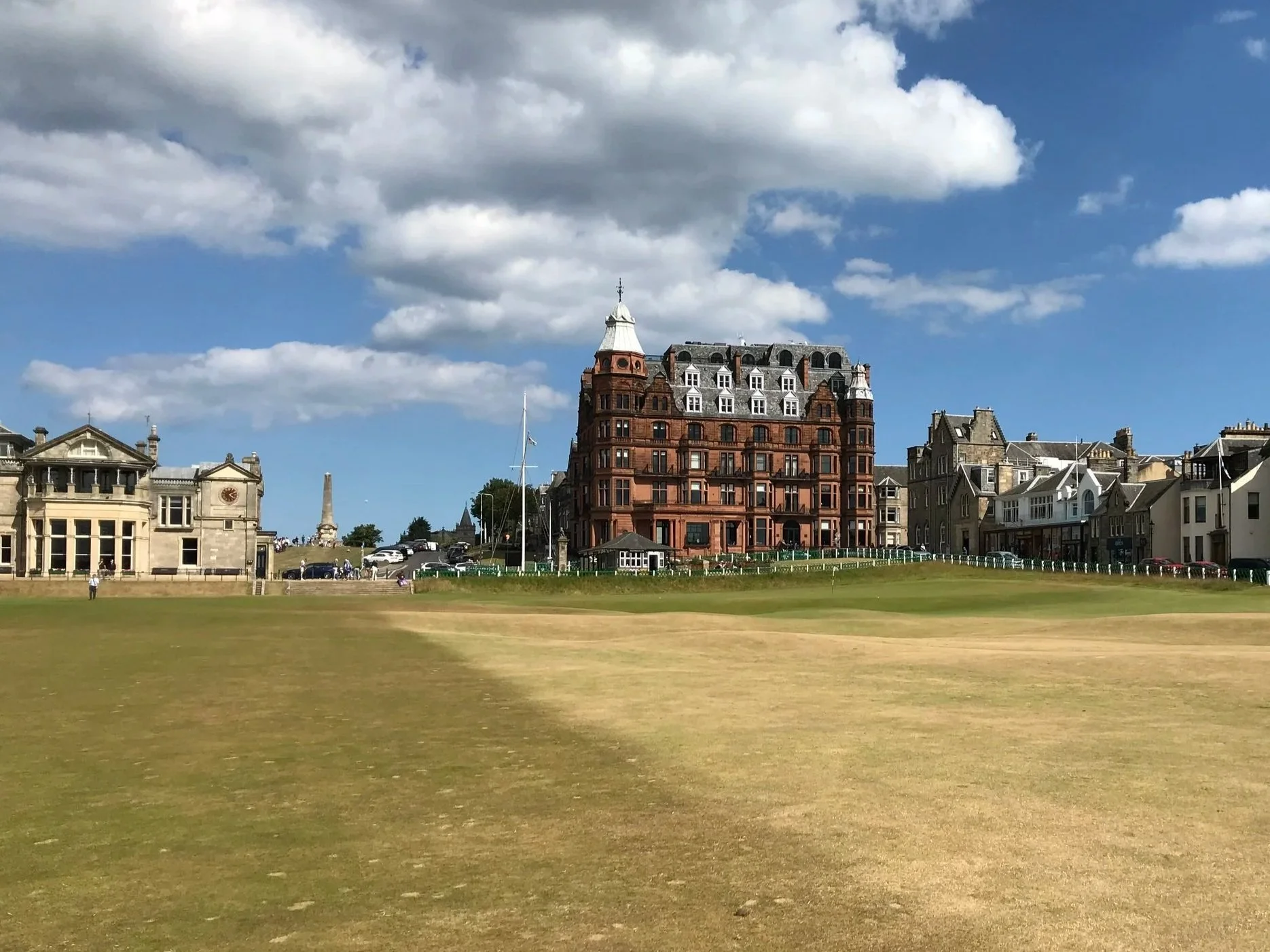 A large grassy area with a historic red-brick building and surrounding architecture, under a partly cloudy blue sky.